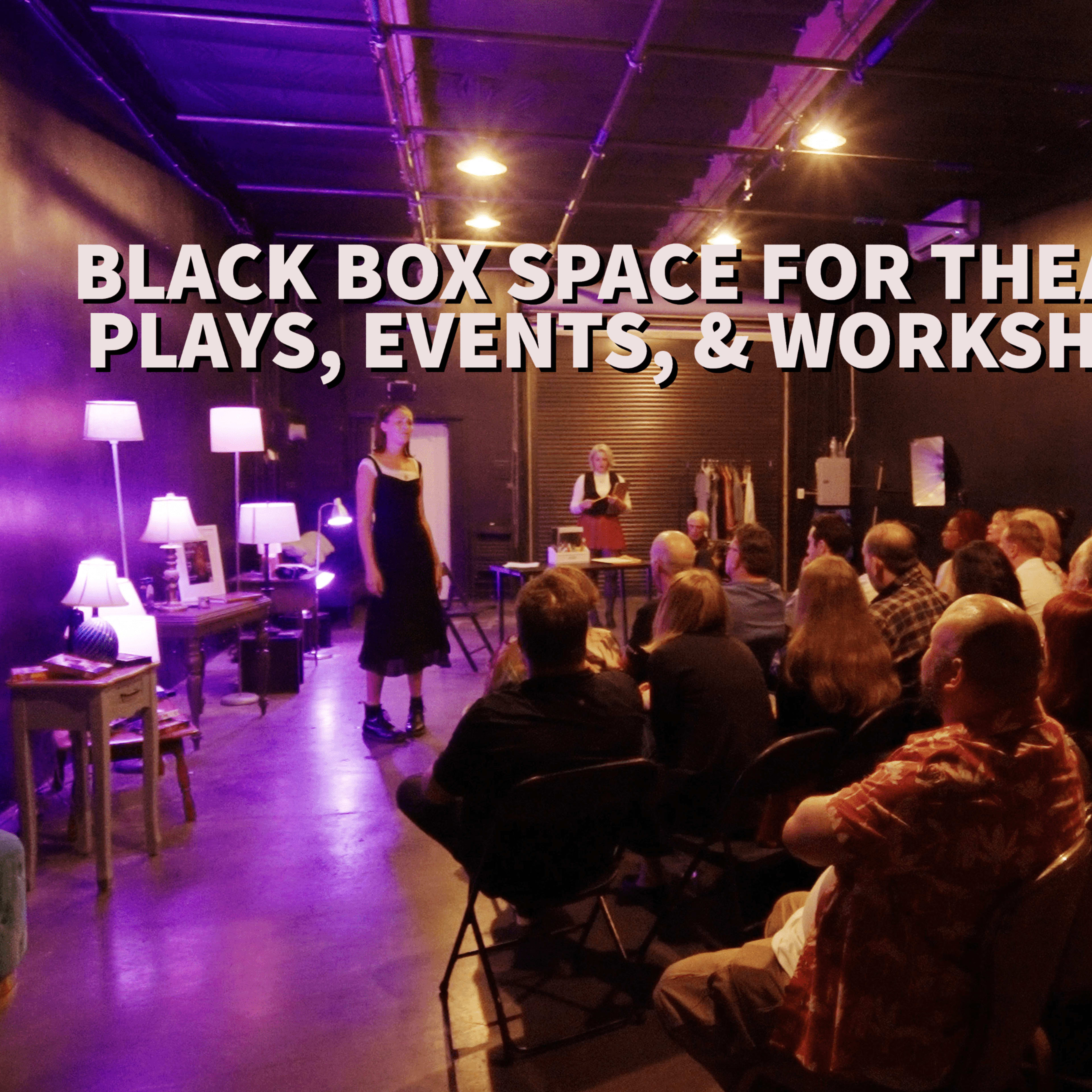 A black box theater space filled with an audience watching a performance, with purple lighting illuminating the stage area.