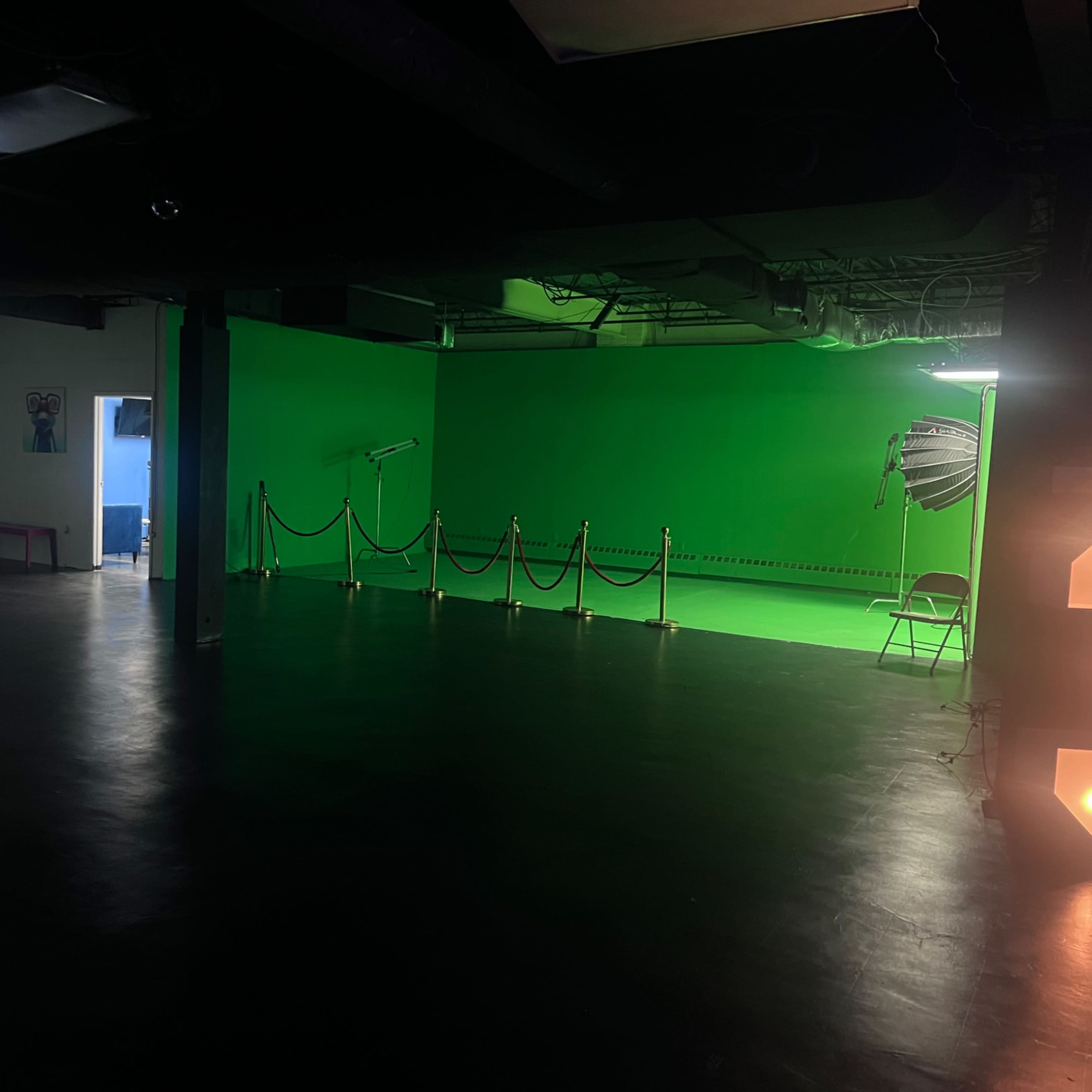 An indoor space featuring a green screen area, a wall displaying a video, and a section with ropes and a chair for audience or production use.
