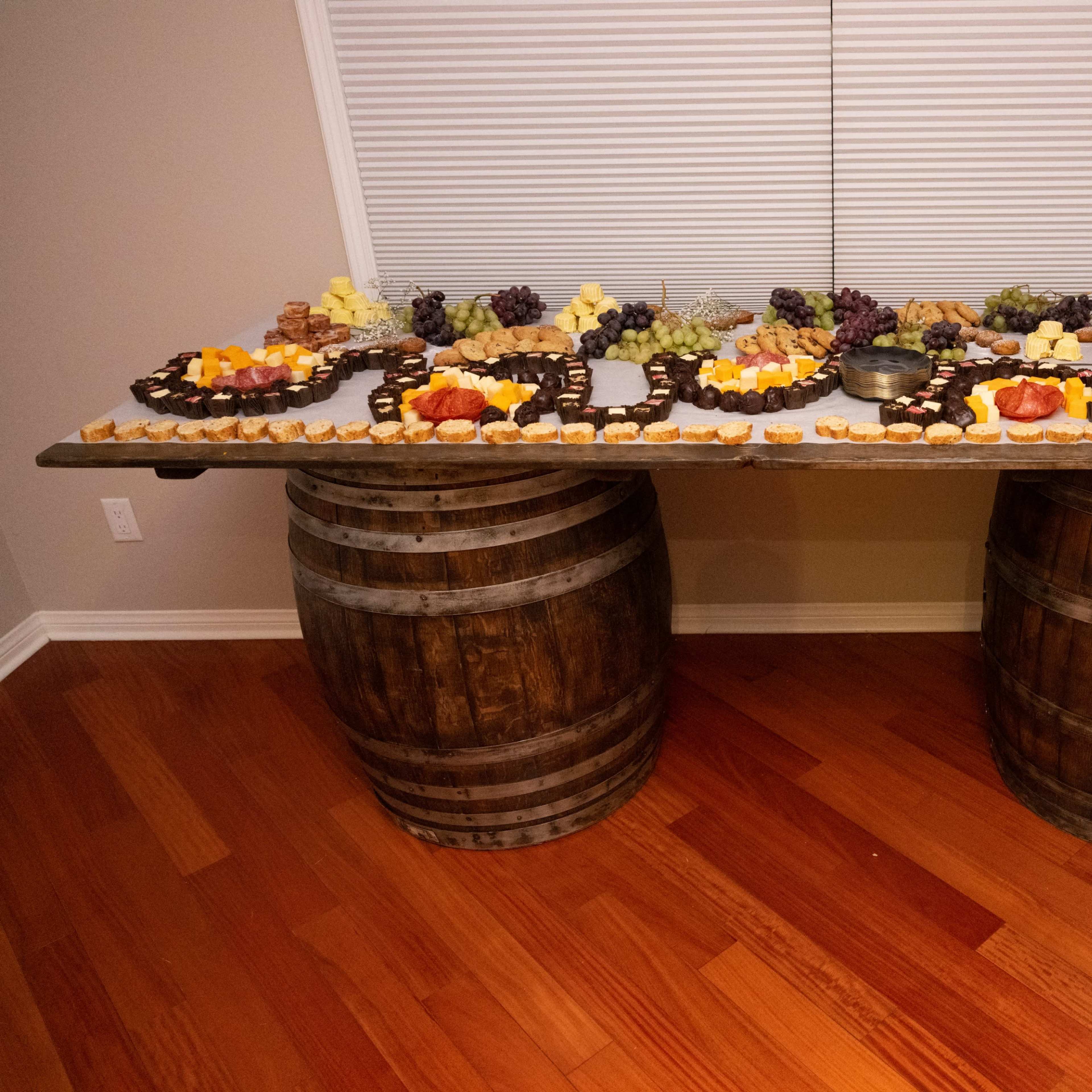 A large, multi-tiered display of assorted fruits, cheeses, and desserts is arranged on a table made from wooden barrels.