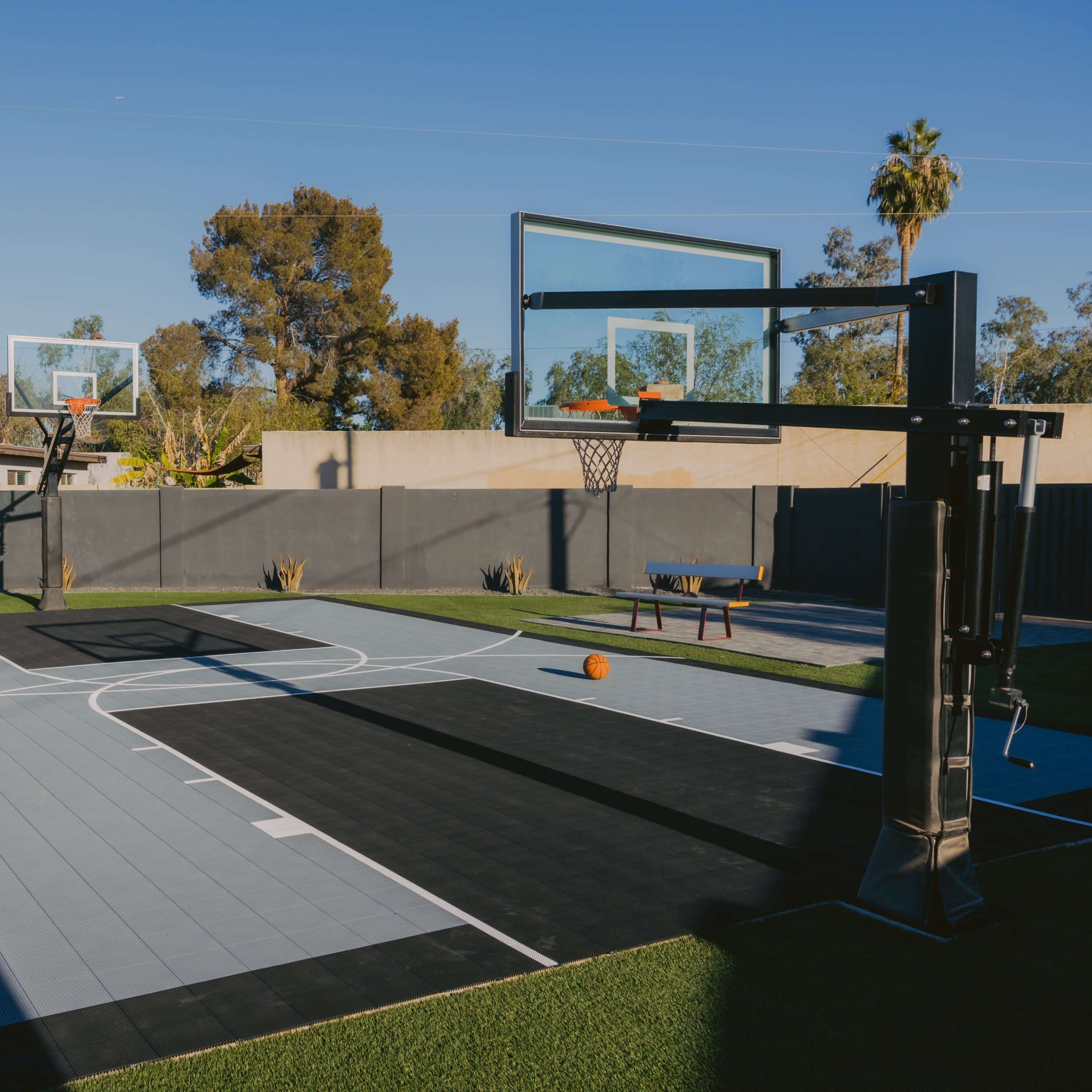 A basketball court with two hoops, surrounded by greenery and a clear blue sky.