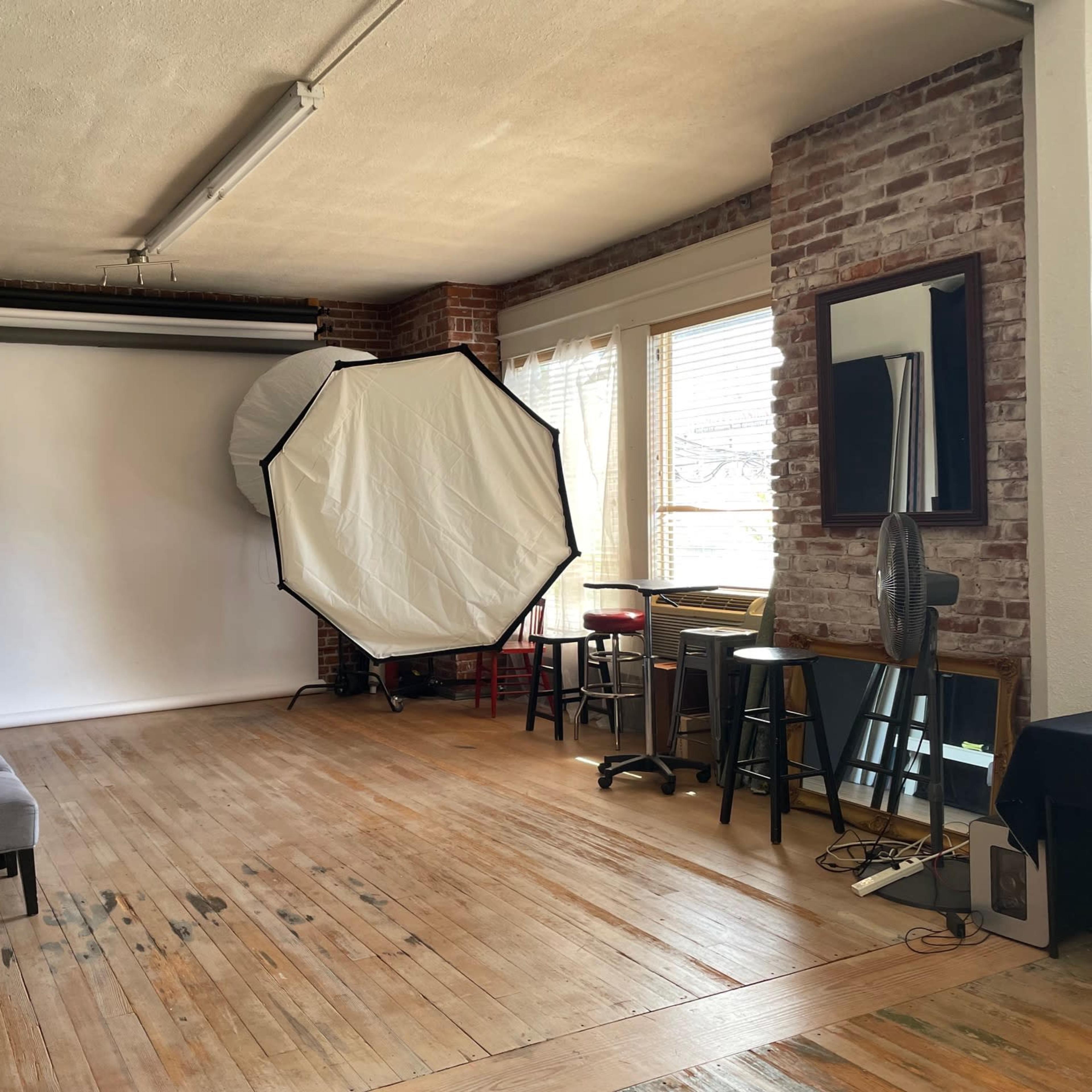 A spacious photography studio with brick walls, wooden flooring, and a backdrop stand featuring a large white screen.