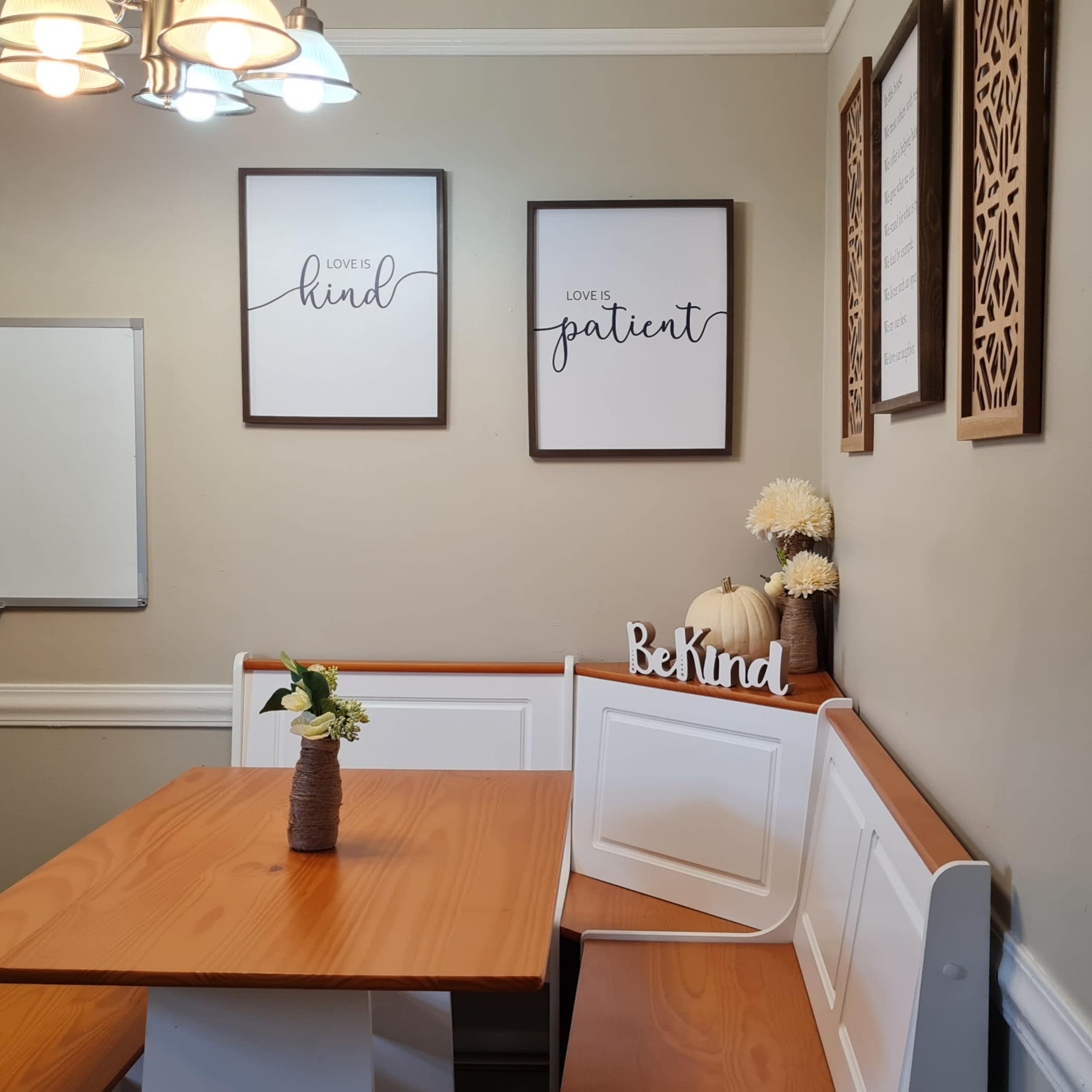 A small dining area with a wooden table, bench seating, framed quotes on the wall, and a whiteboard nearby.