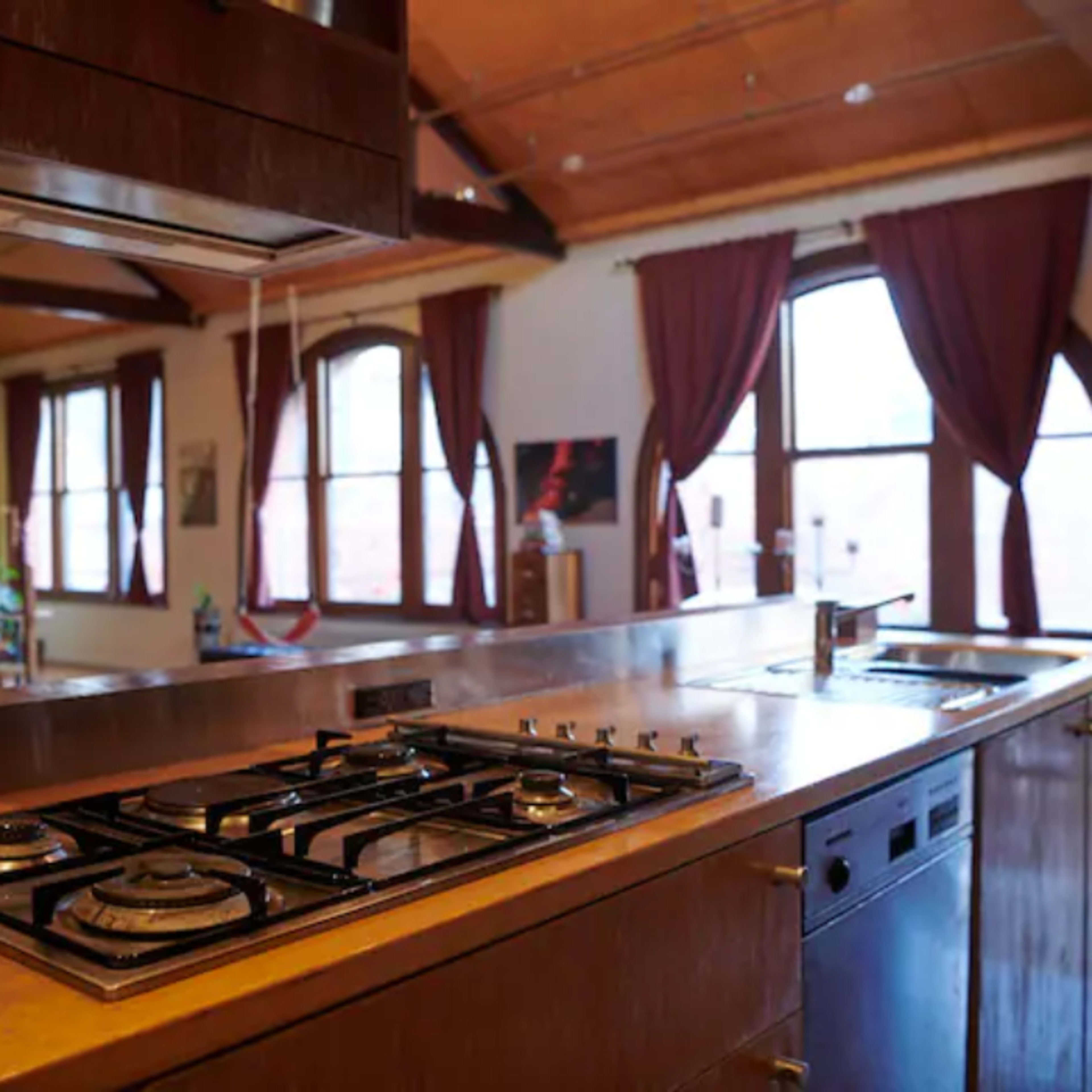 A modern kitchen with a gas stove, a stainless steel countertop, and large windows framed by curtains.