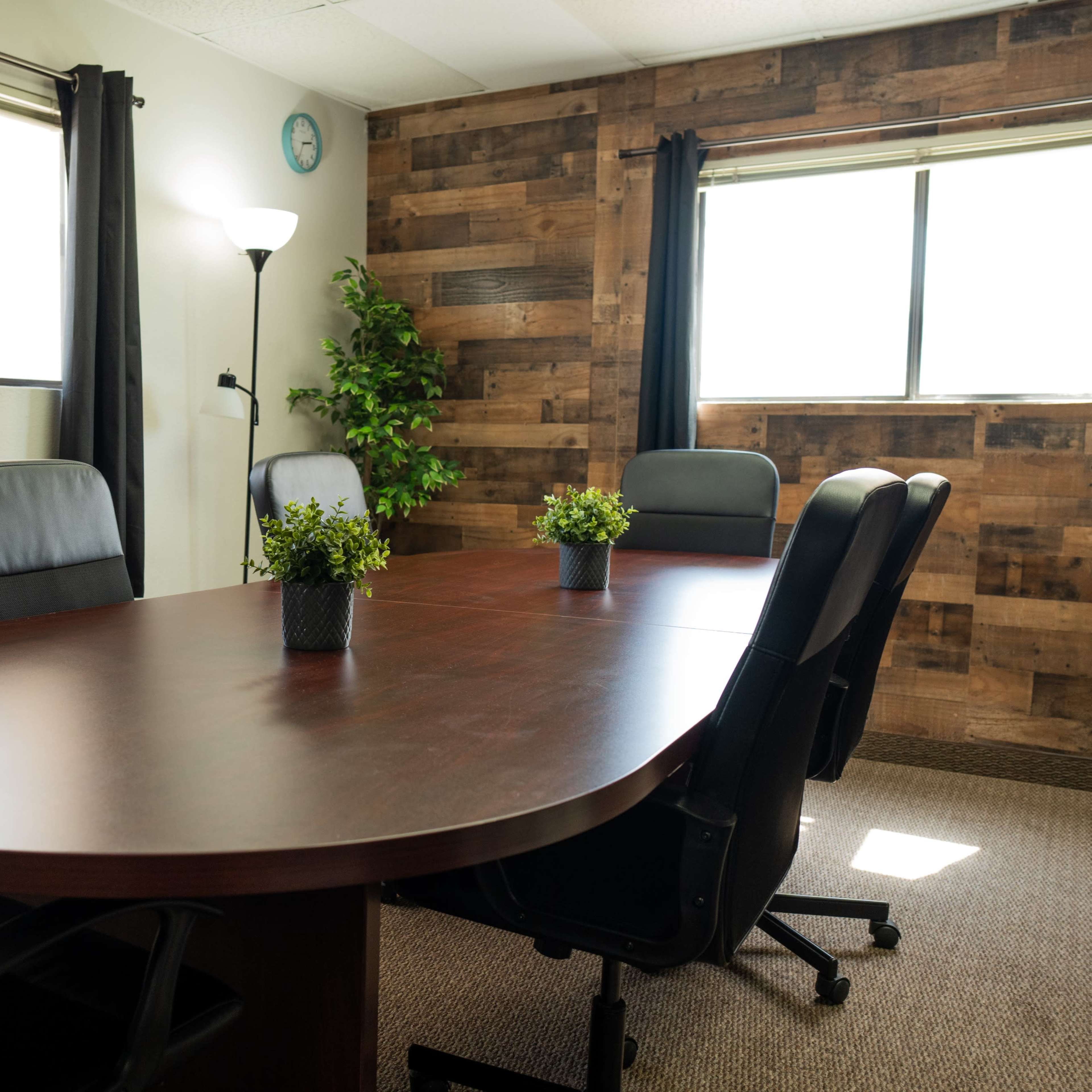 A conference room features a large oval wooden table surrounded by black office chairs, with large windows and a wooden accent wall.