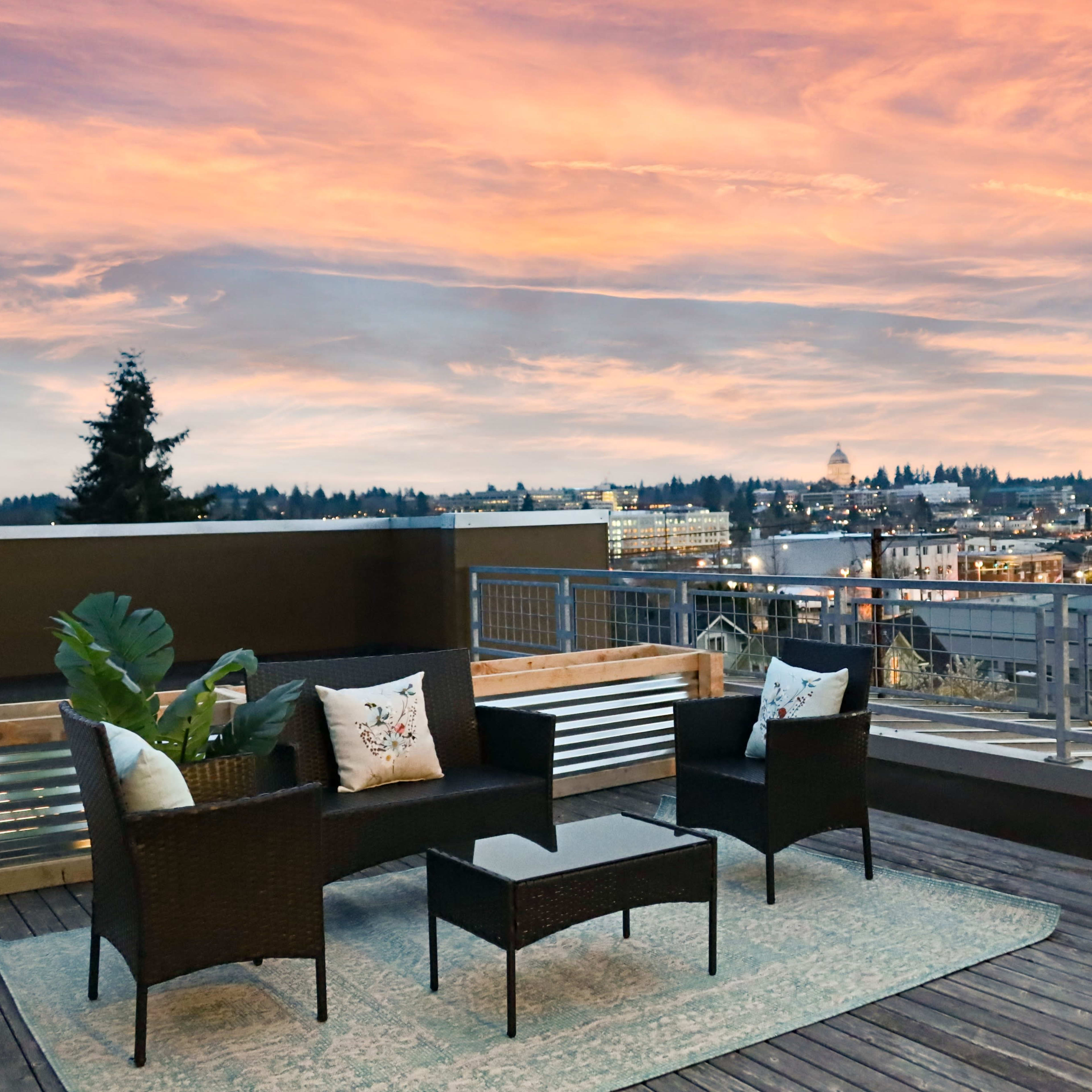 A rooftop terrace features a set of outdoor furniture, including two chairs and a coffee table, overlooking a cityscape at sunset.