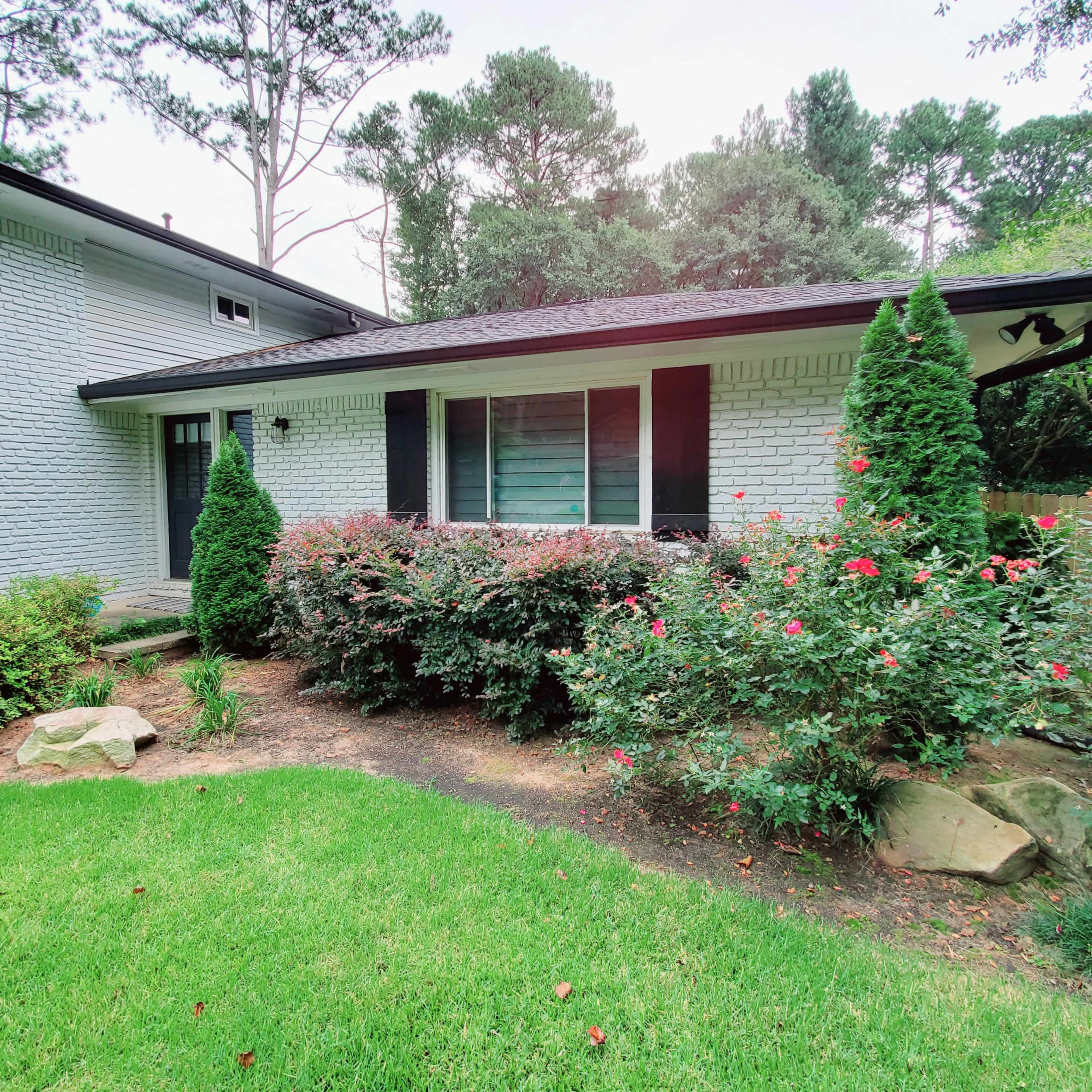 A brick house with a porch surrounded by neatly trimmed shrubs and rose bushes on a grassy lawn.