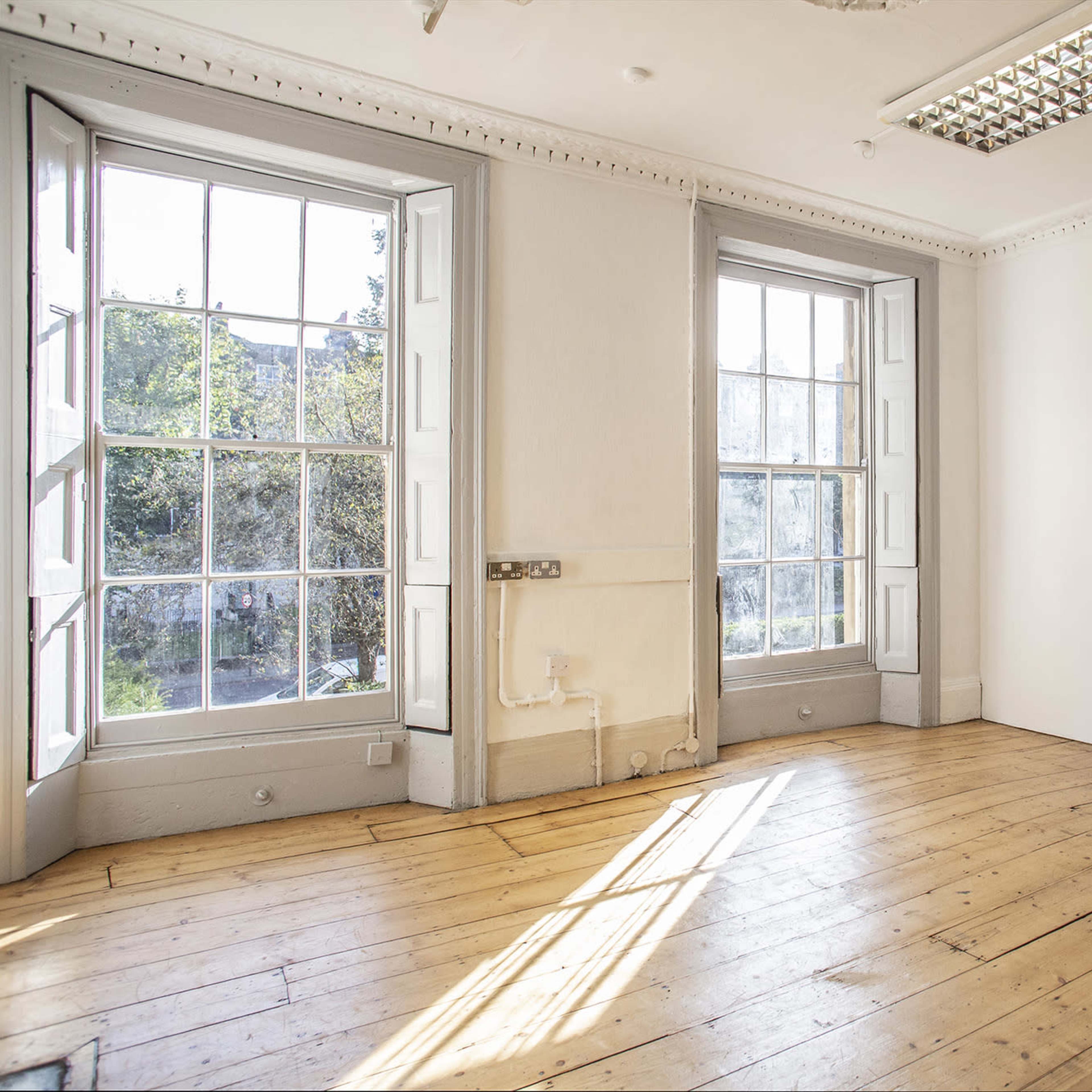 A bright, empty room features two large windows with open shutters, exposing wooden floors and a plain white wall.