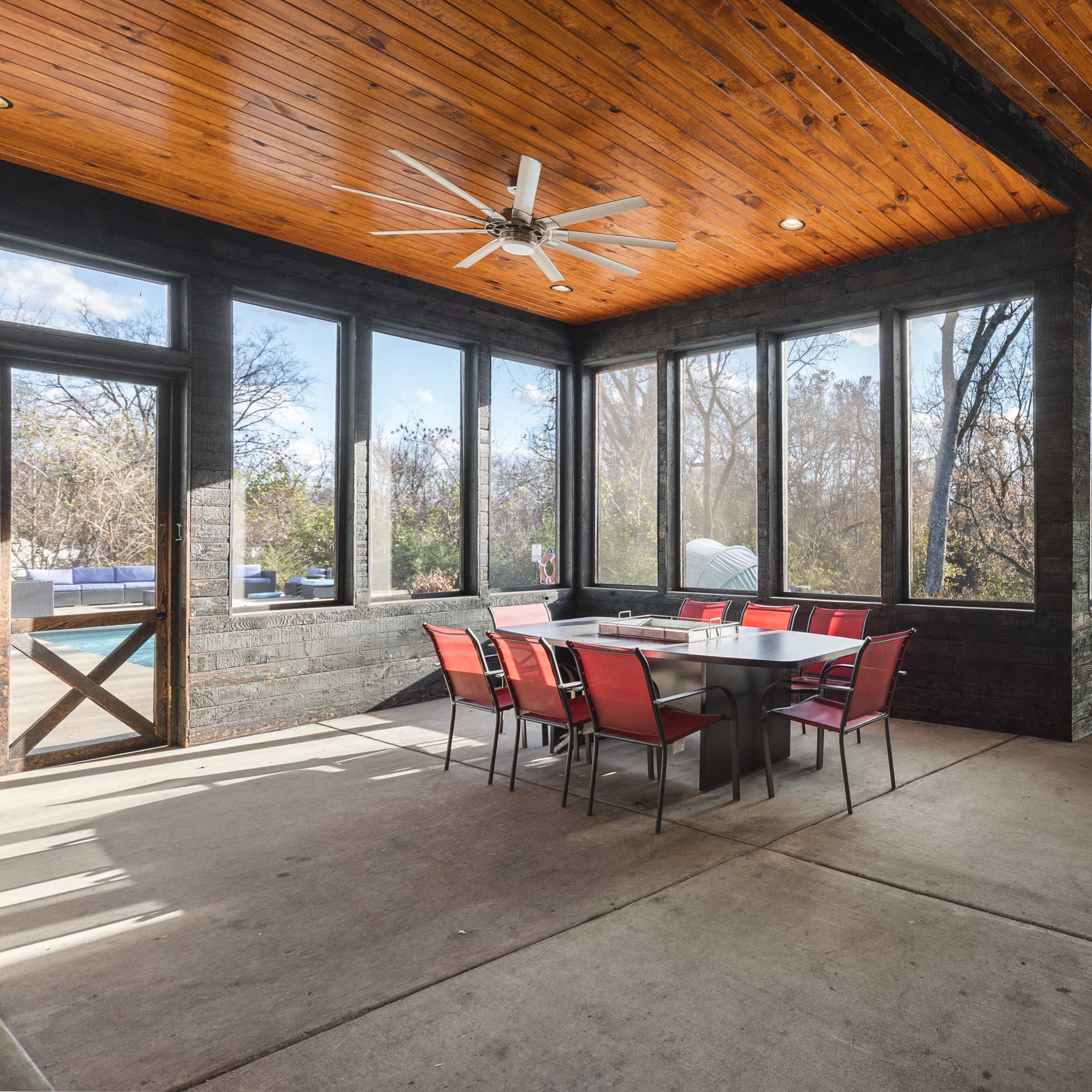 A screened-in porch featuring a large dining table with red chairs, wooden ceiling beams, and a motorcycle parked in one corner.