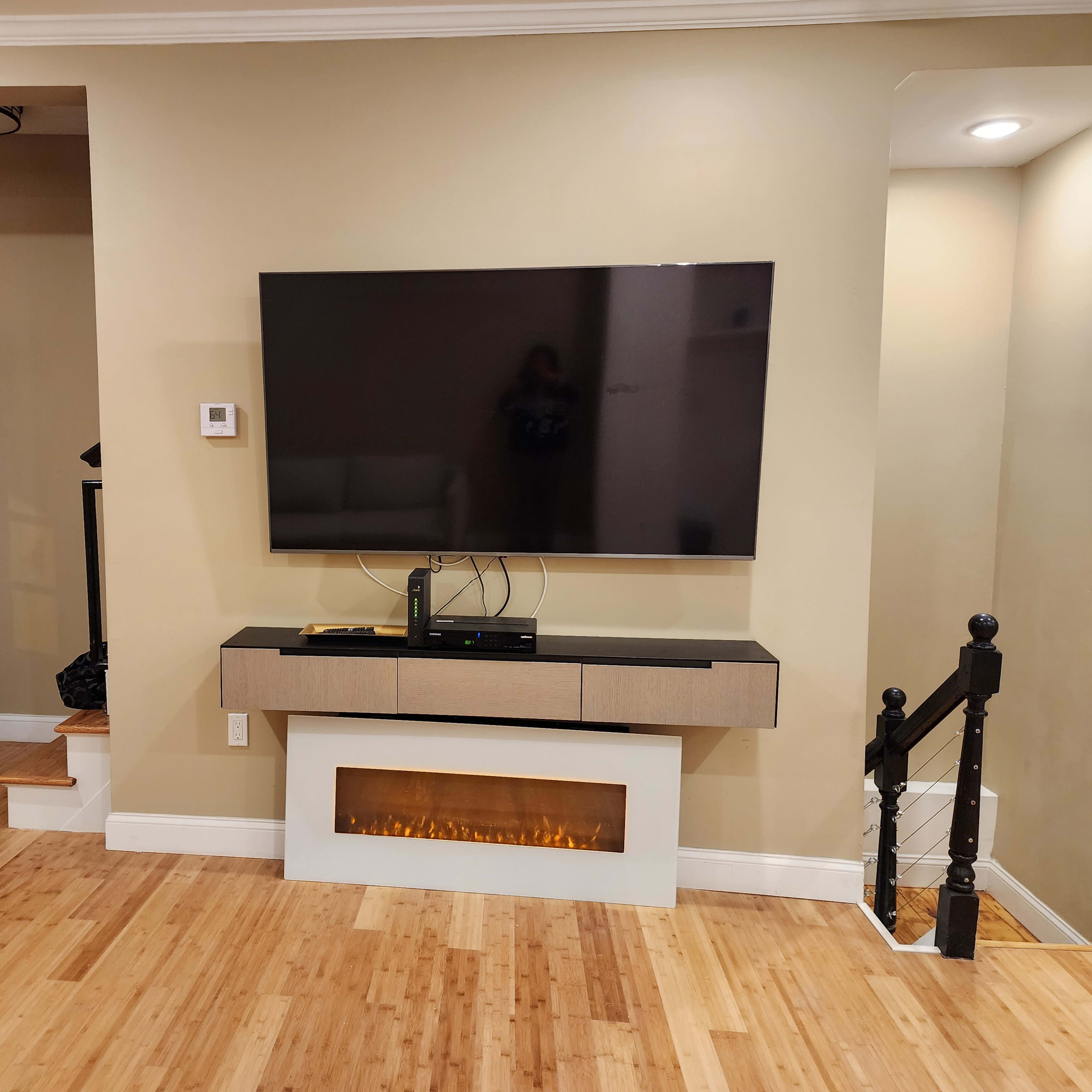 A wall-mounted television is positioned above a sleek console with an electric fireplace, adjacent to a staircase and entrance door in a room with light-colored walls and a bamboo floor.