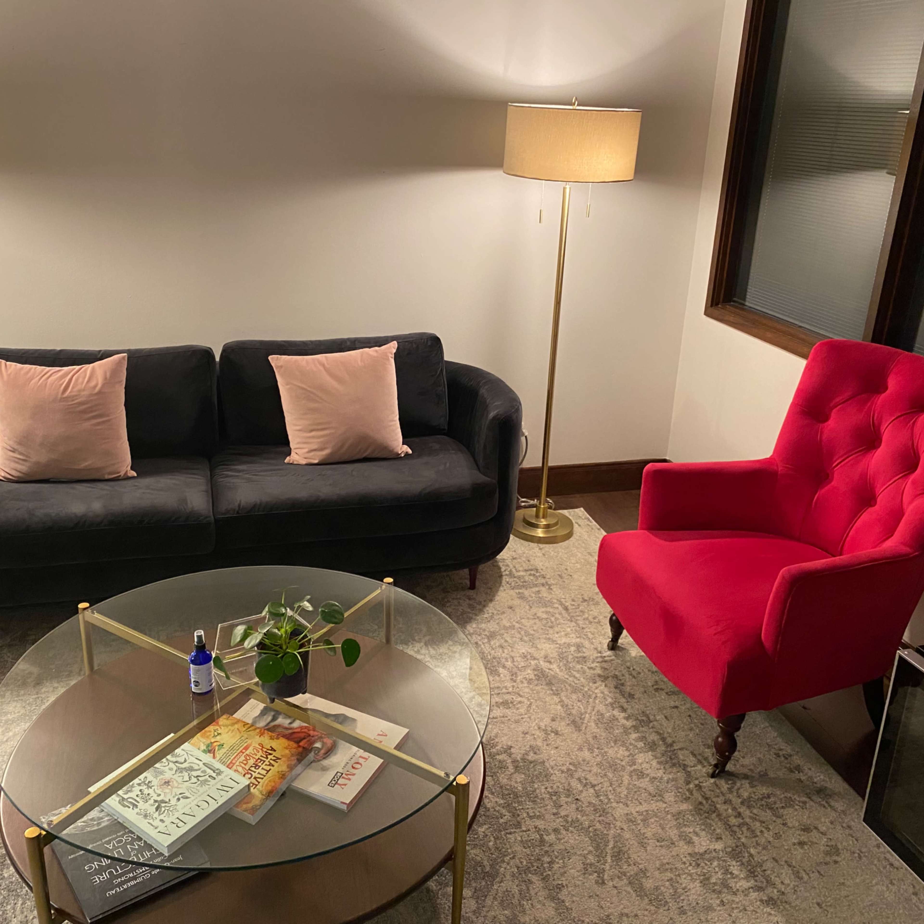 A black sofa with pink cushions faces a red armchair, alongside a round glass coffee table with various magazines and a small plant.