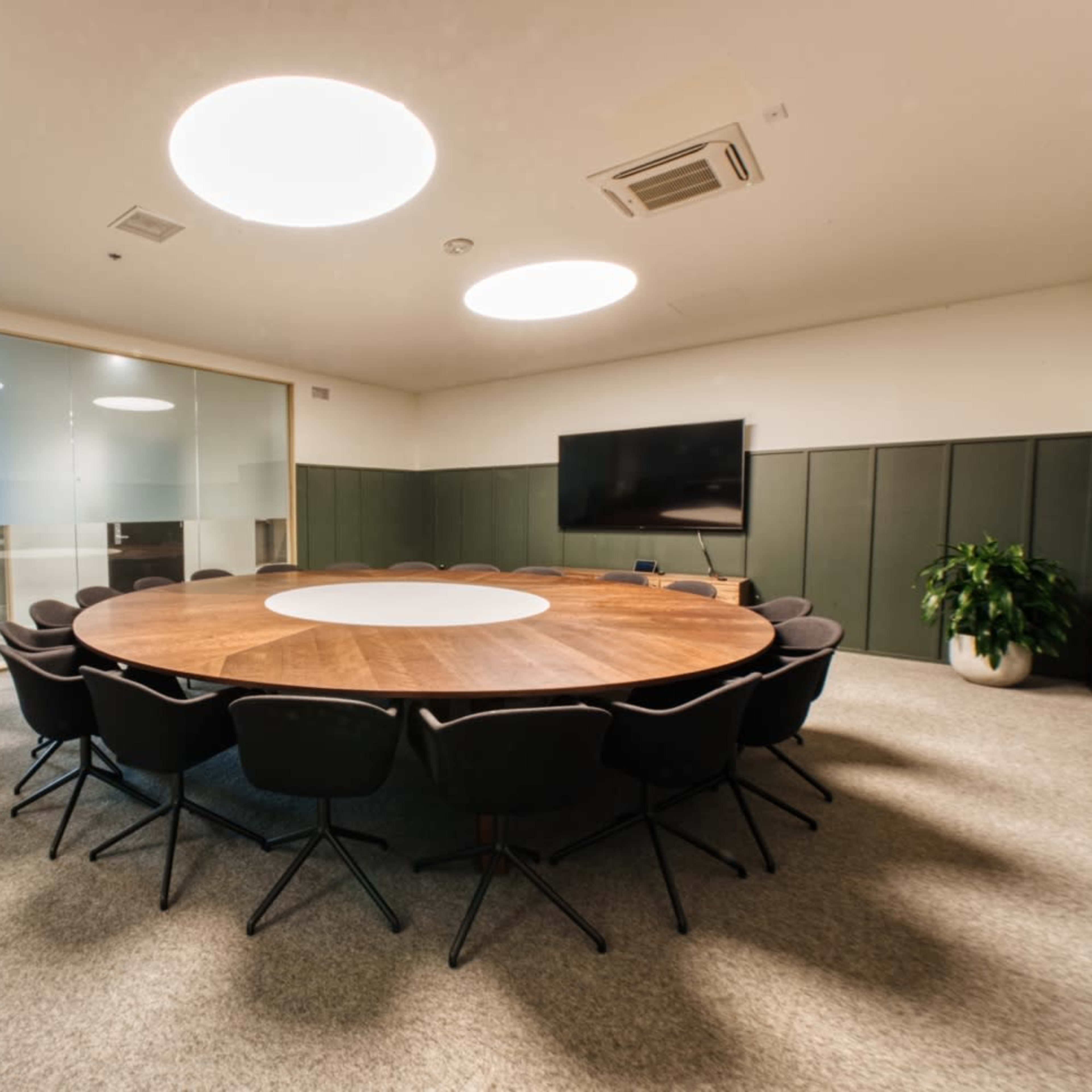 A modern conference room featuring a circular wooden table surrounded by black chairs, with glass walls and a large screen on one side.