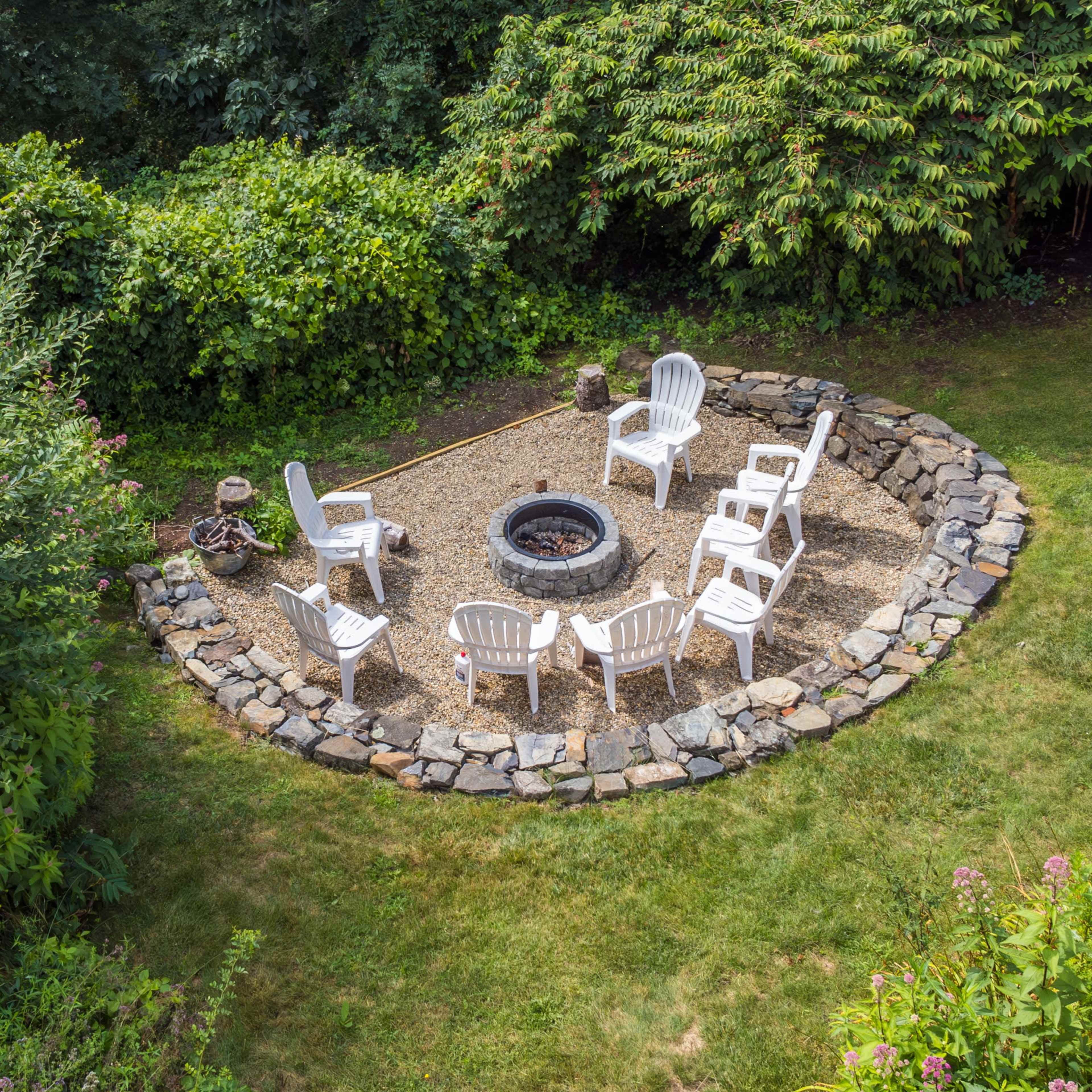 A circular seating area with white chairs surrounds a stone fire pit, nestled among lush greenery.