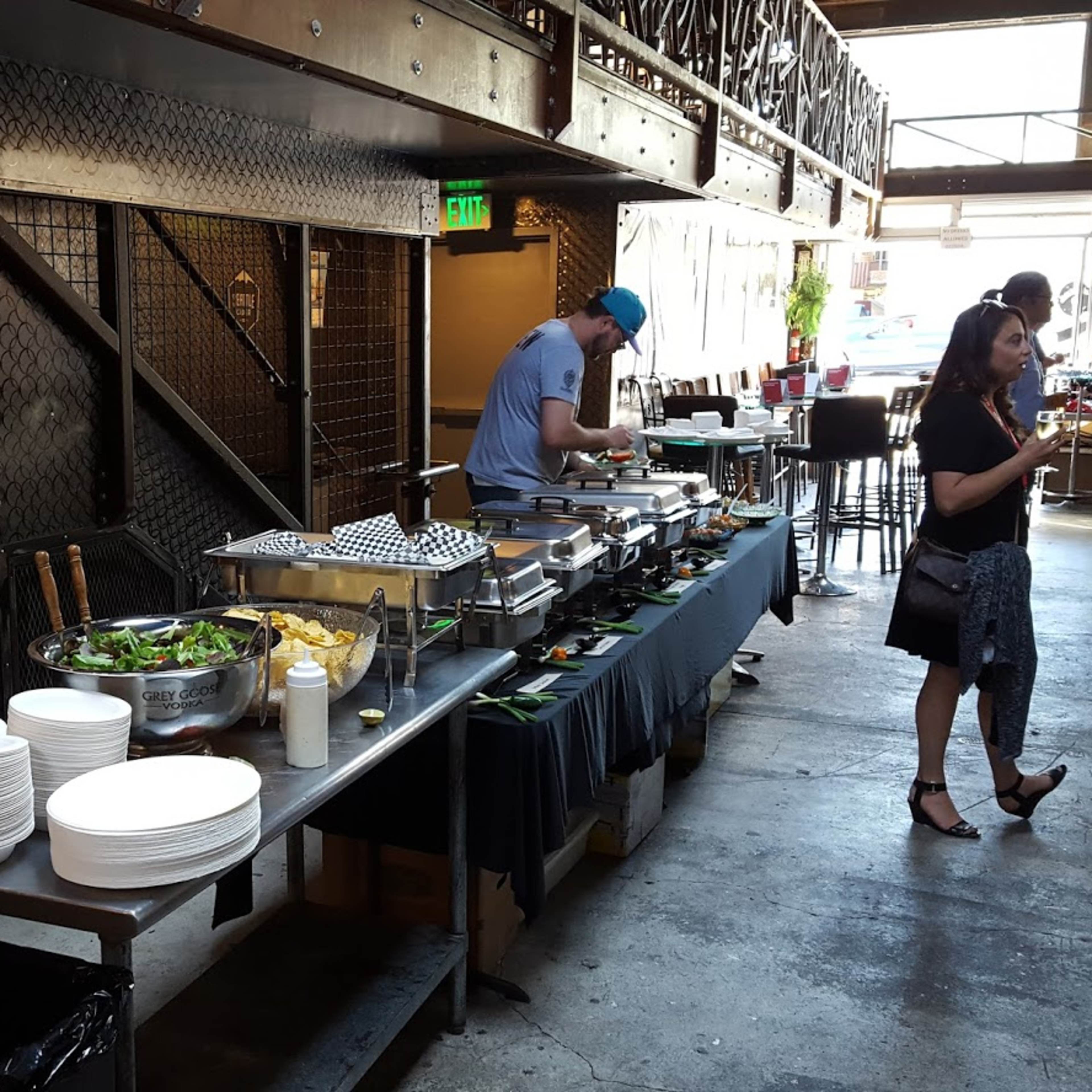 A buffet setup with food trays, plates, and a salad station is arranged along a metal table in a venue featuring an industrial design.