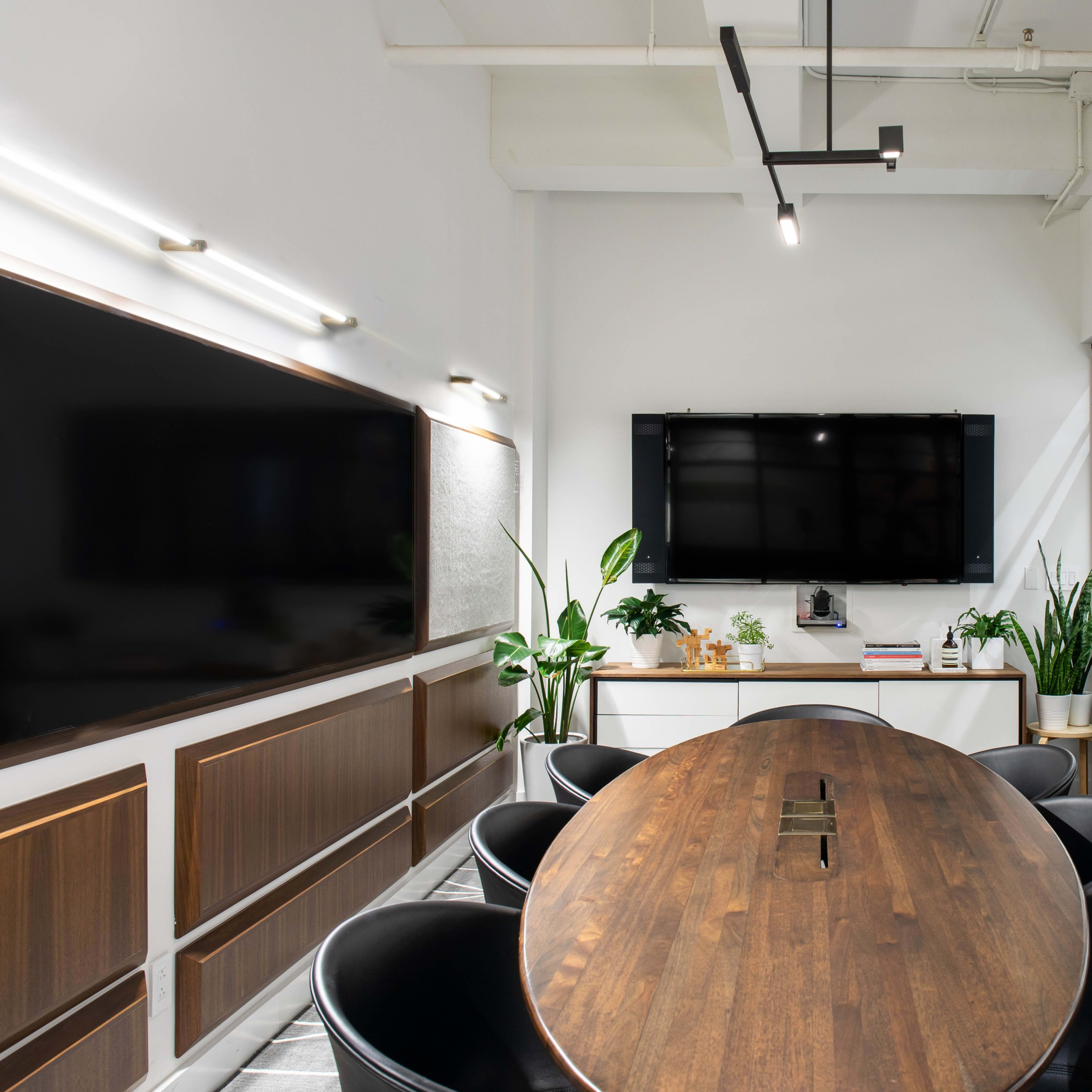 A modern conference room features a large wooden oval table surrounded by black chairs, with two wall-mounted screens and plants in the background.