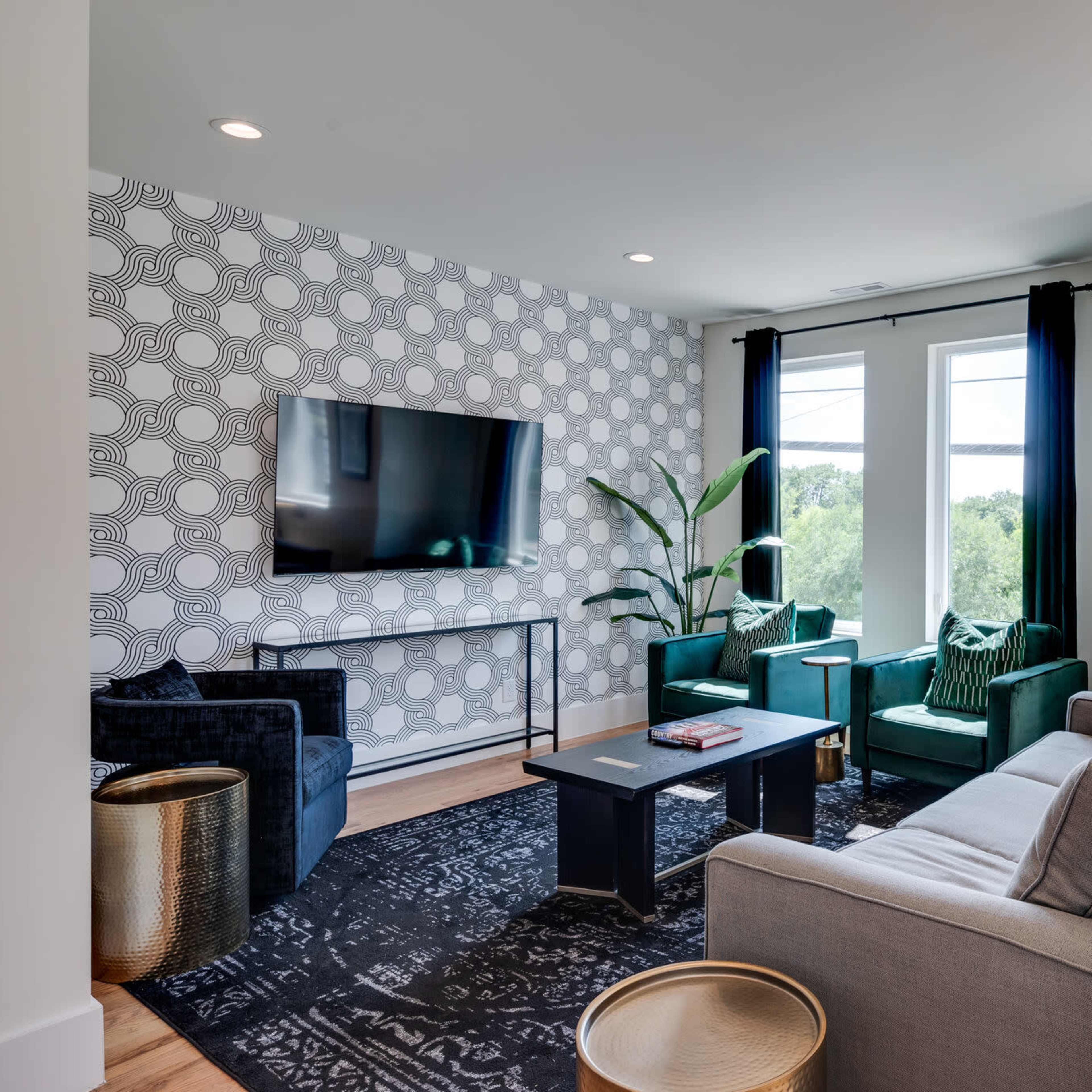 A modern living room featuring a flat-screen TV mounted on a patterned wall, green armchairs, a gray sofa, and large windows providing natural light.