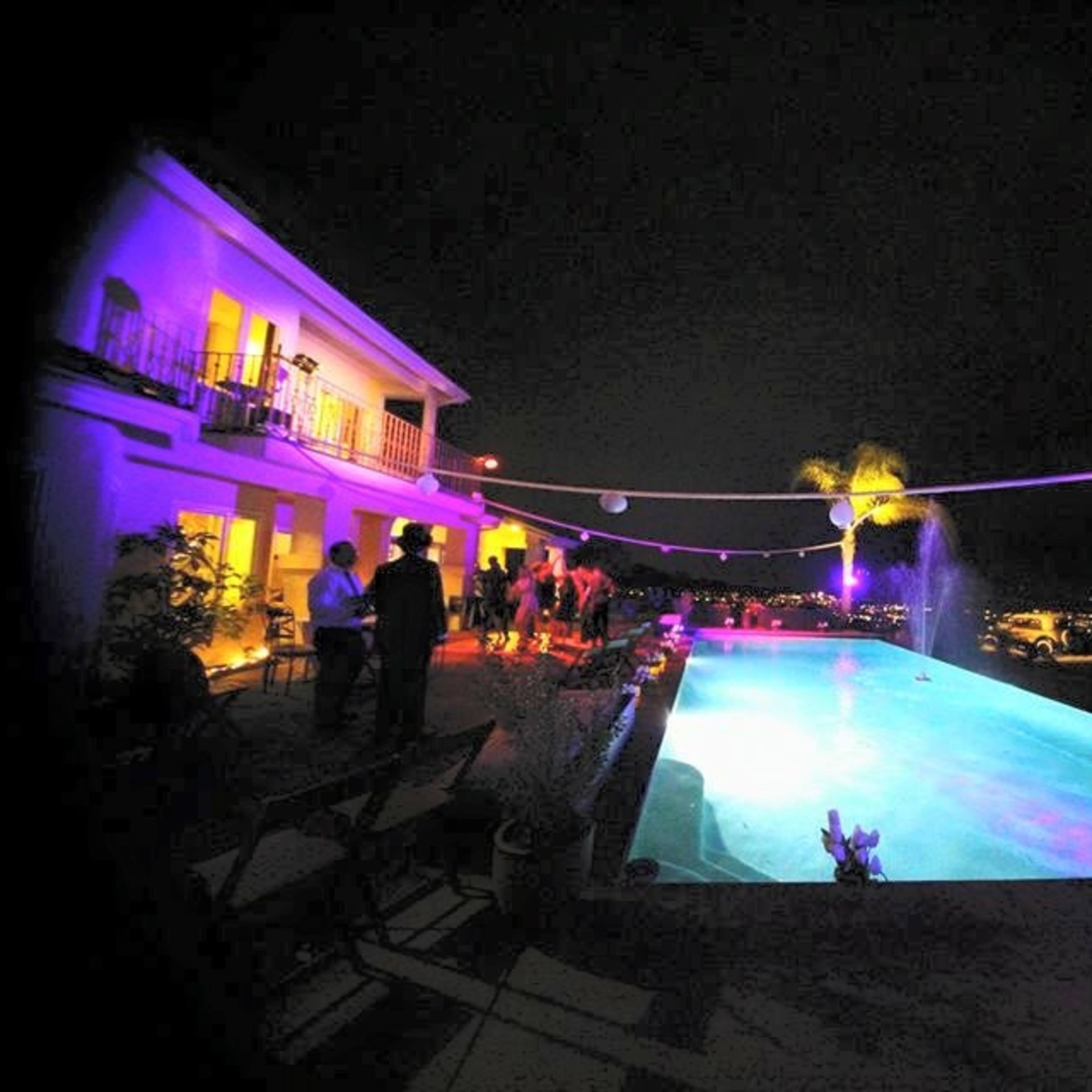 A well-lit outdoor pool area is filled with guests enjoying an evening gathering, with city lights visible in the background.