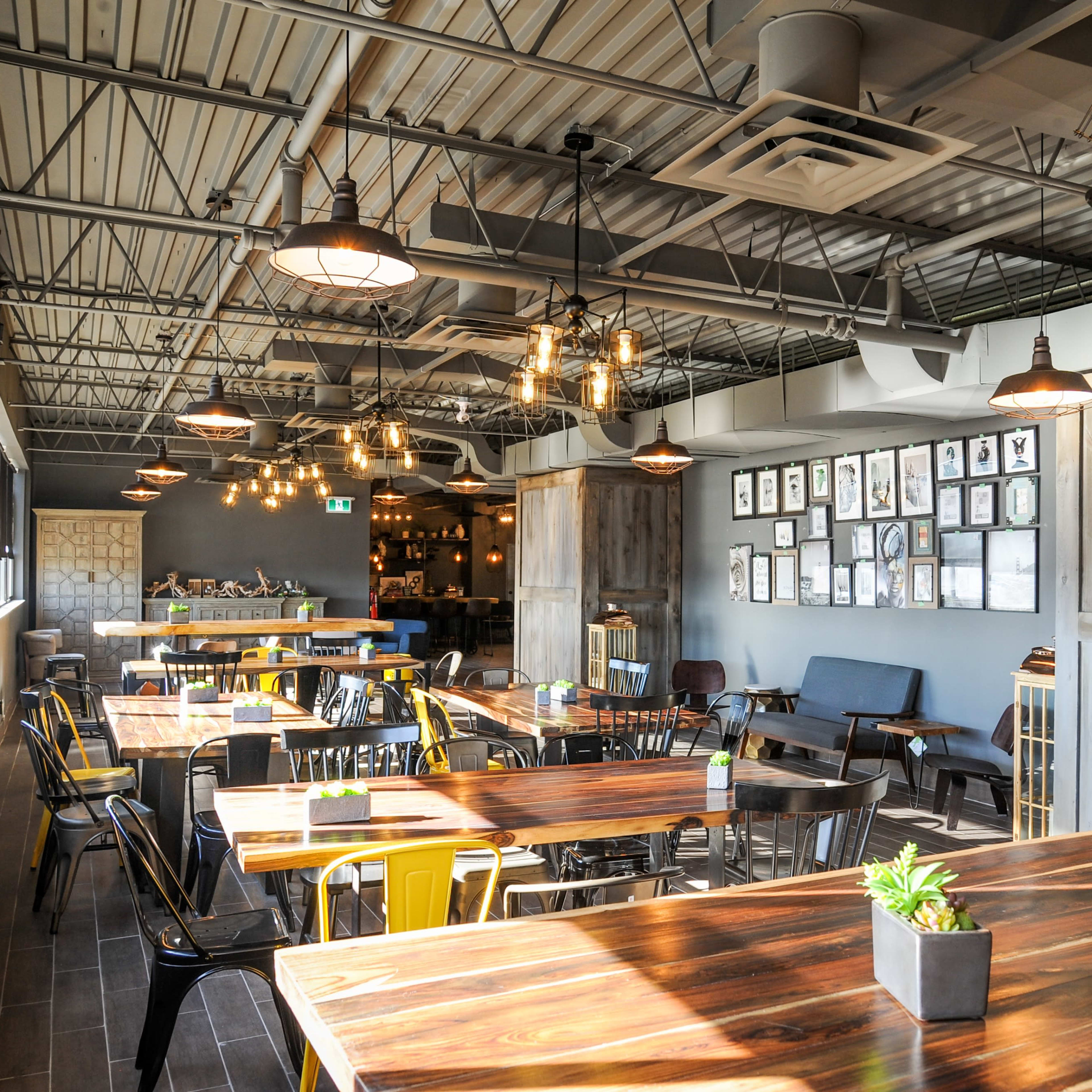 A spacious restaurant interior with wooden tables, black and yellow chairs, and various framed artworks on the walls, illuminated by pendant lights.