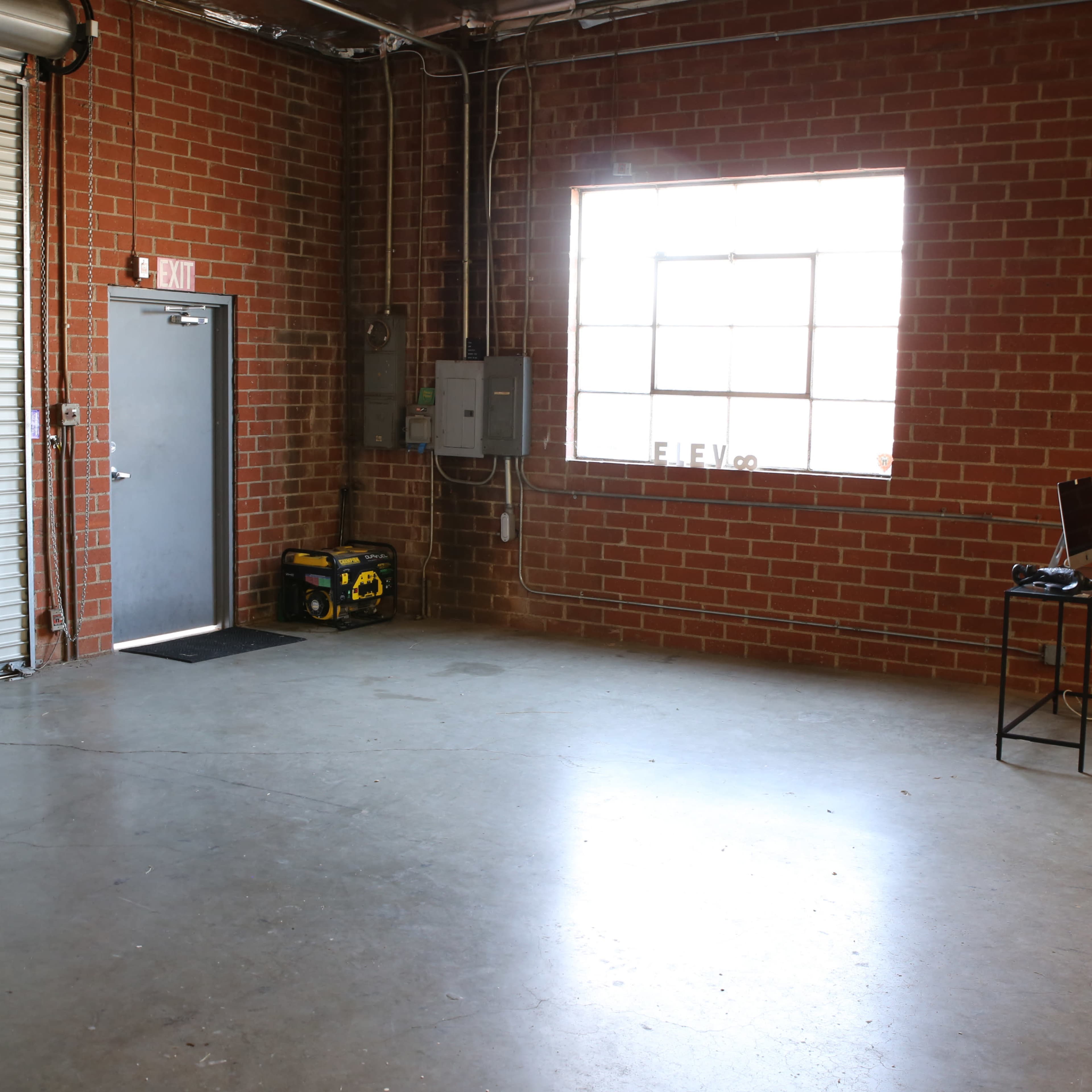 An empty industrial room with a large window, a metal door, and a chair beside a small table.