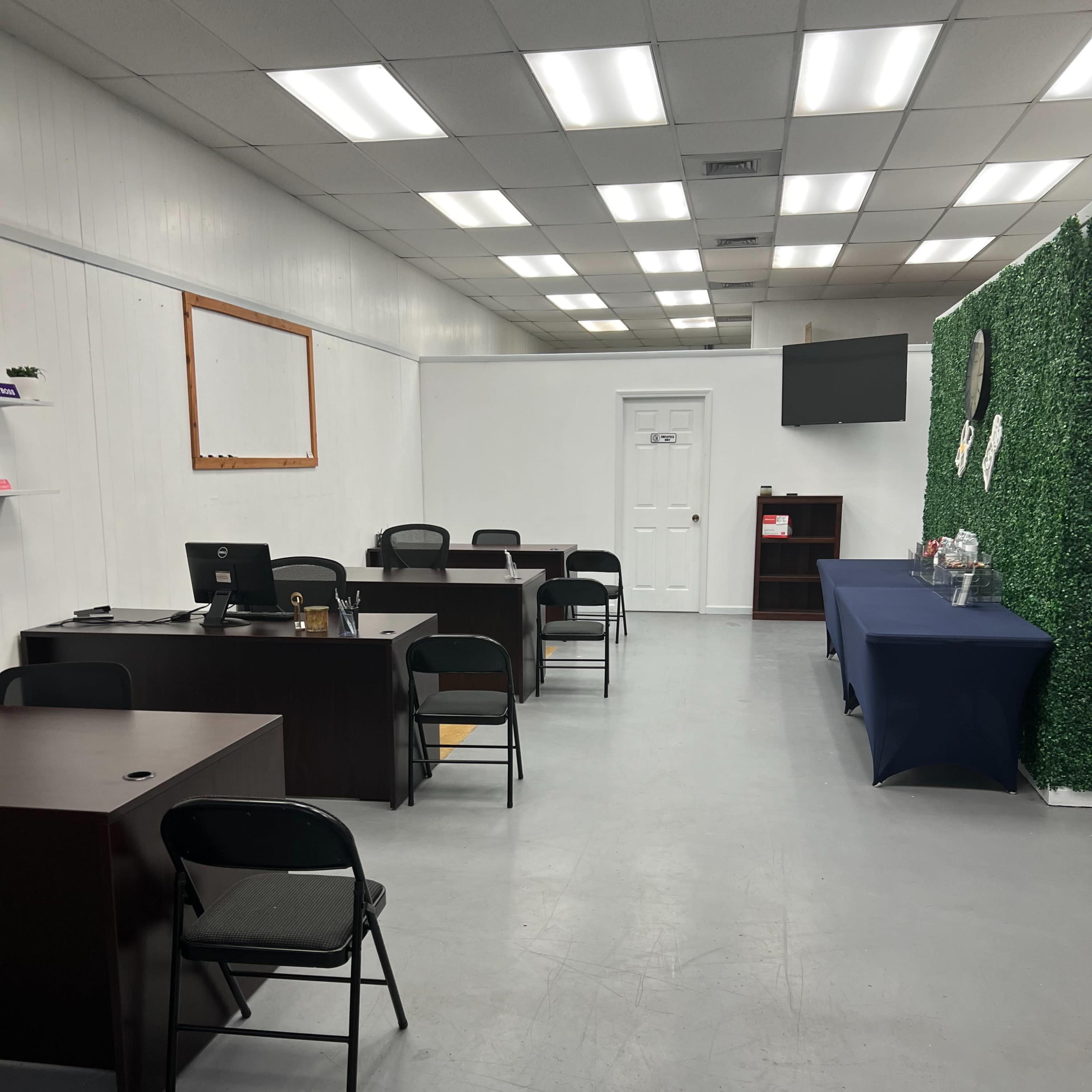 An office space with several black chairs and desks arranged in two rows, along with a table covered with a blue cloth and a wall decorated with green foliage.