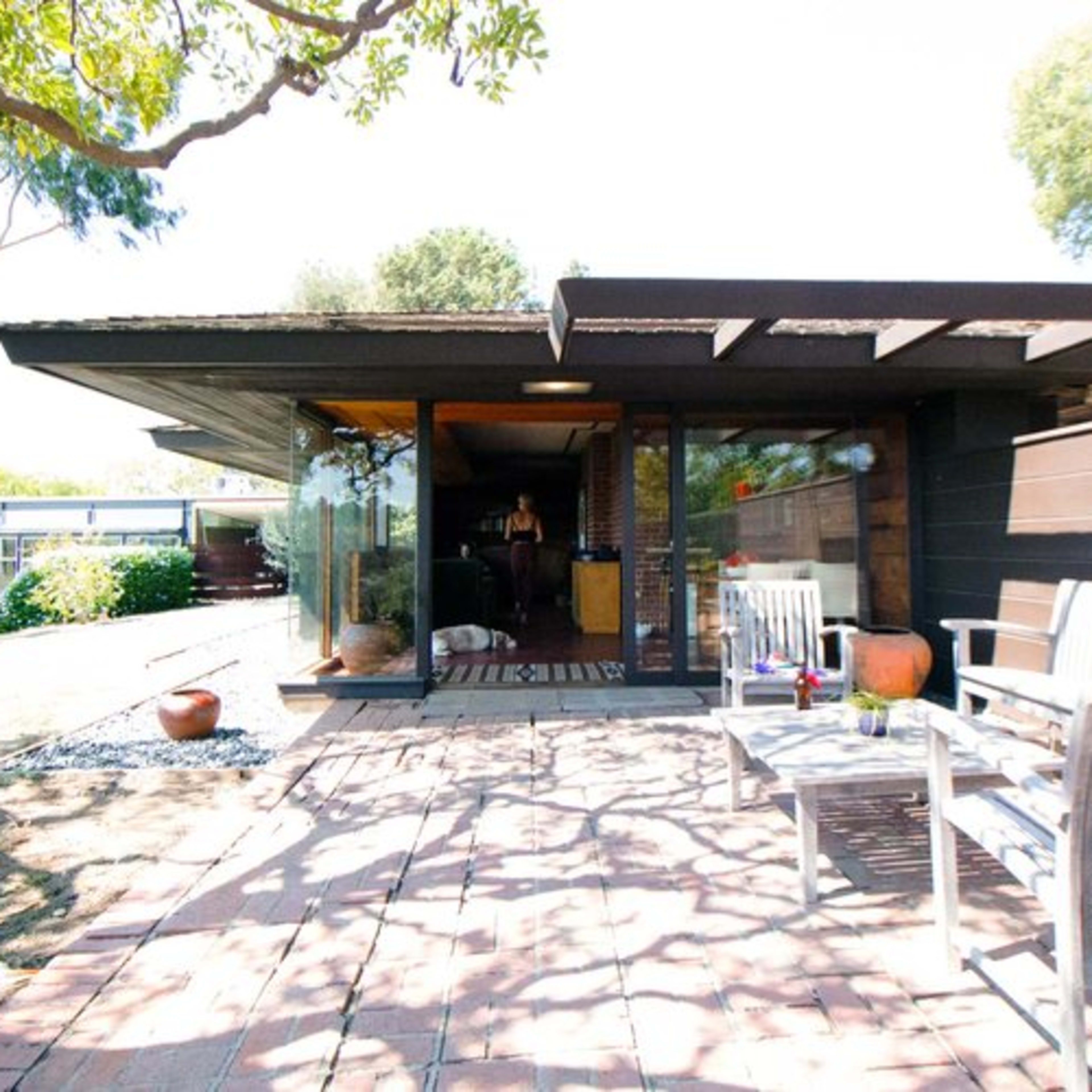 A modern house with a flat roof and large glass doors opens to a patio surrounded by trees and a well-maintained garden.