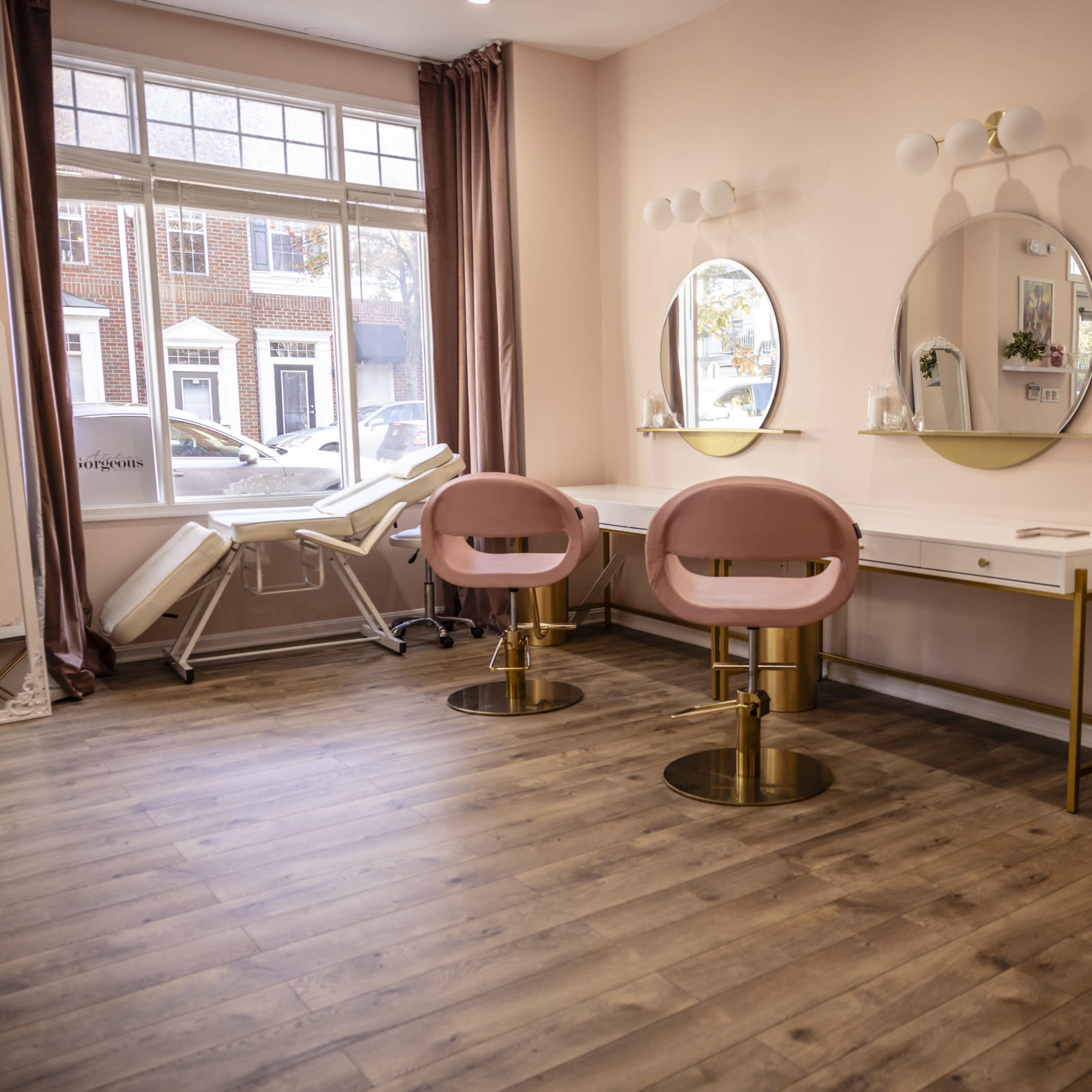 A beauty salon interior with three round mirrors and pink chairs arranged around white dressing tables under natural light from large windows.