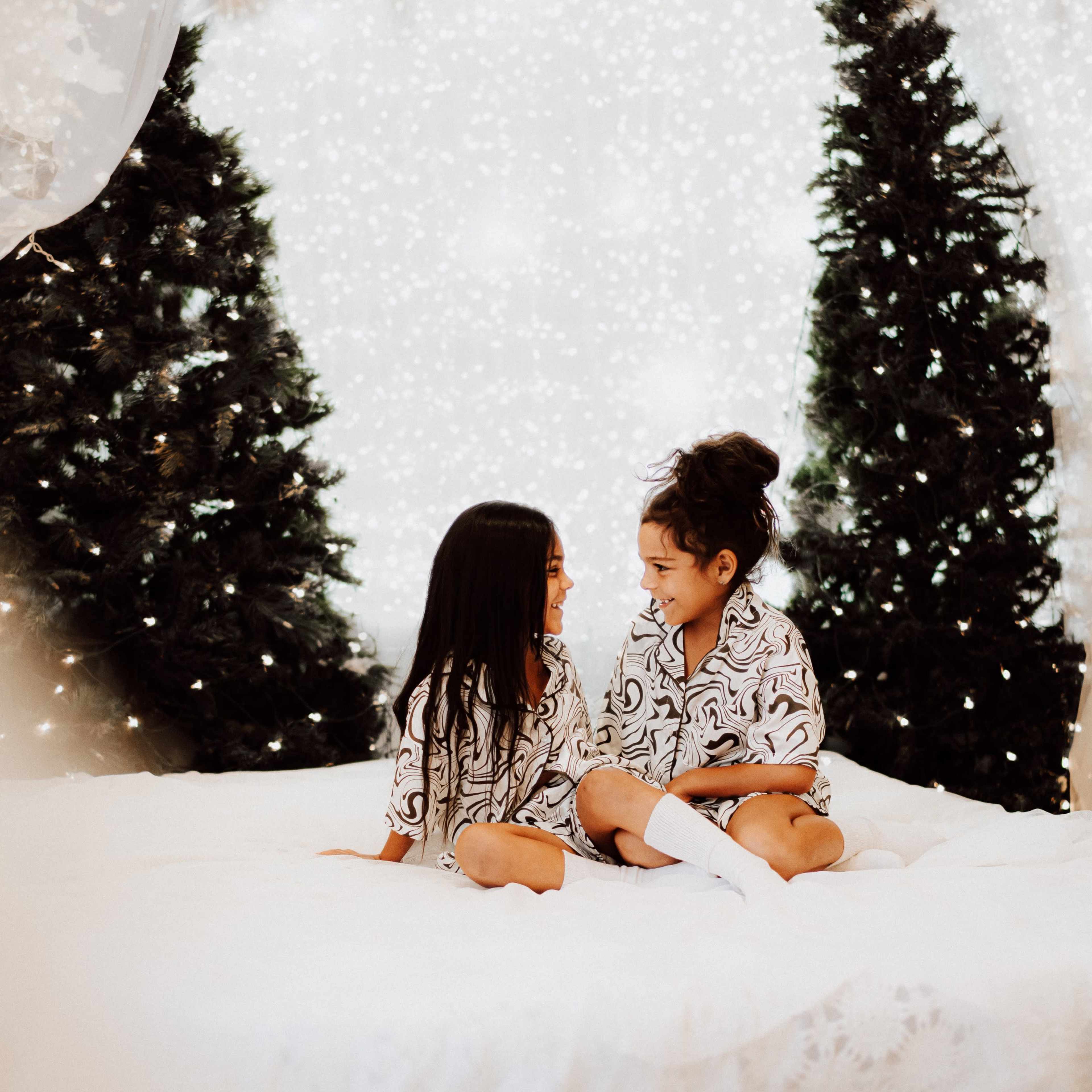 Two children sit on a white bed surrounded by decorated Christmas trees and a backdrop of soft white lights.