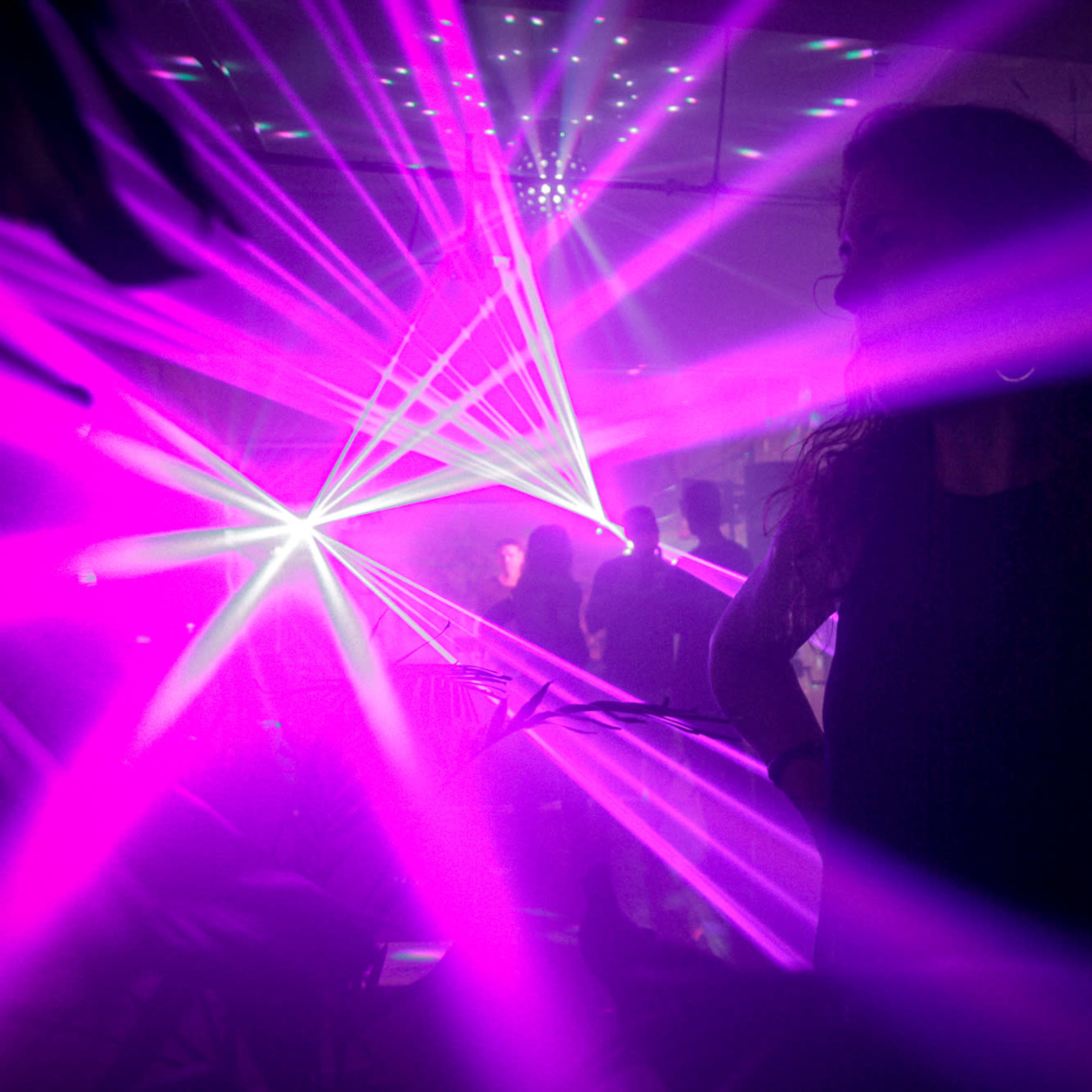 A woman stands with her hands on her hips in a dimly lit room filled with purple laser lights and silhouettes of people dancing.