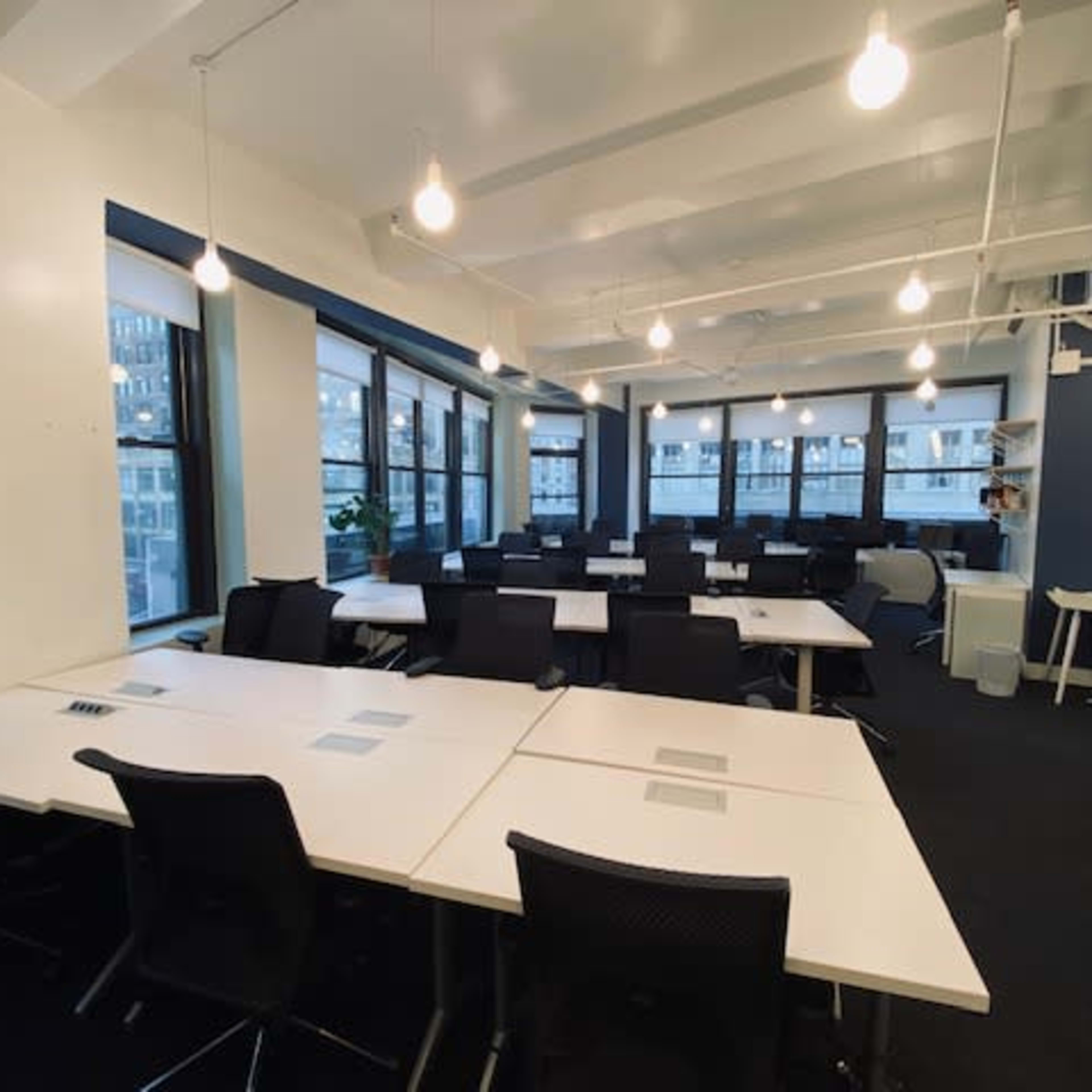 A modern office space with multiple empty desks arranged in rows, ample natural light coming through large windows, and overhead lighting.