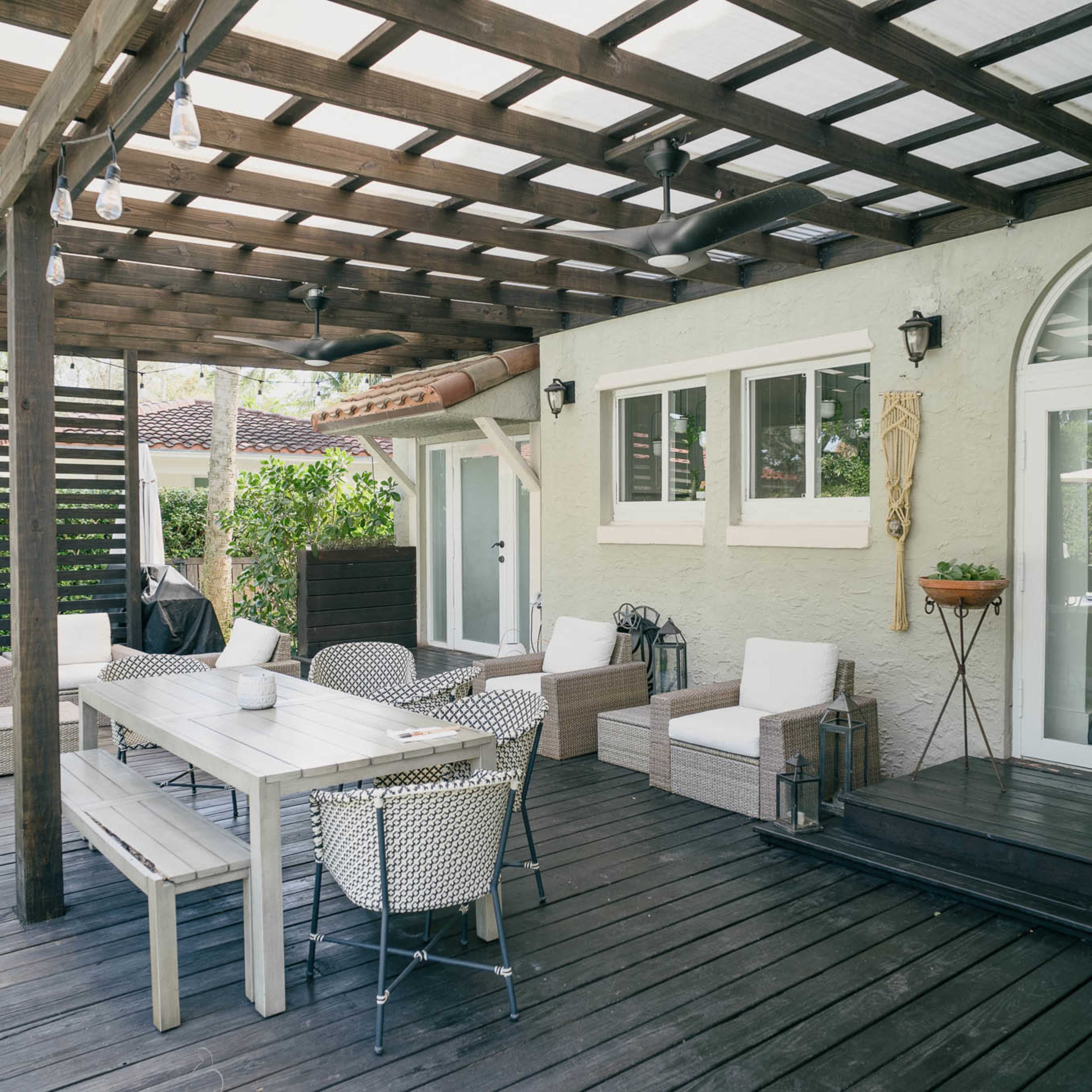A covered wooden patio with a large dining table, seating areas, and well-maintained greenery in the background.