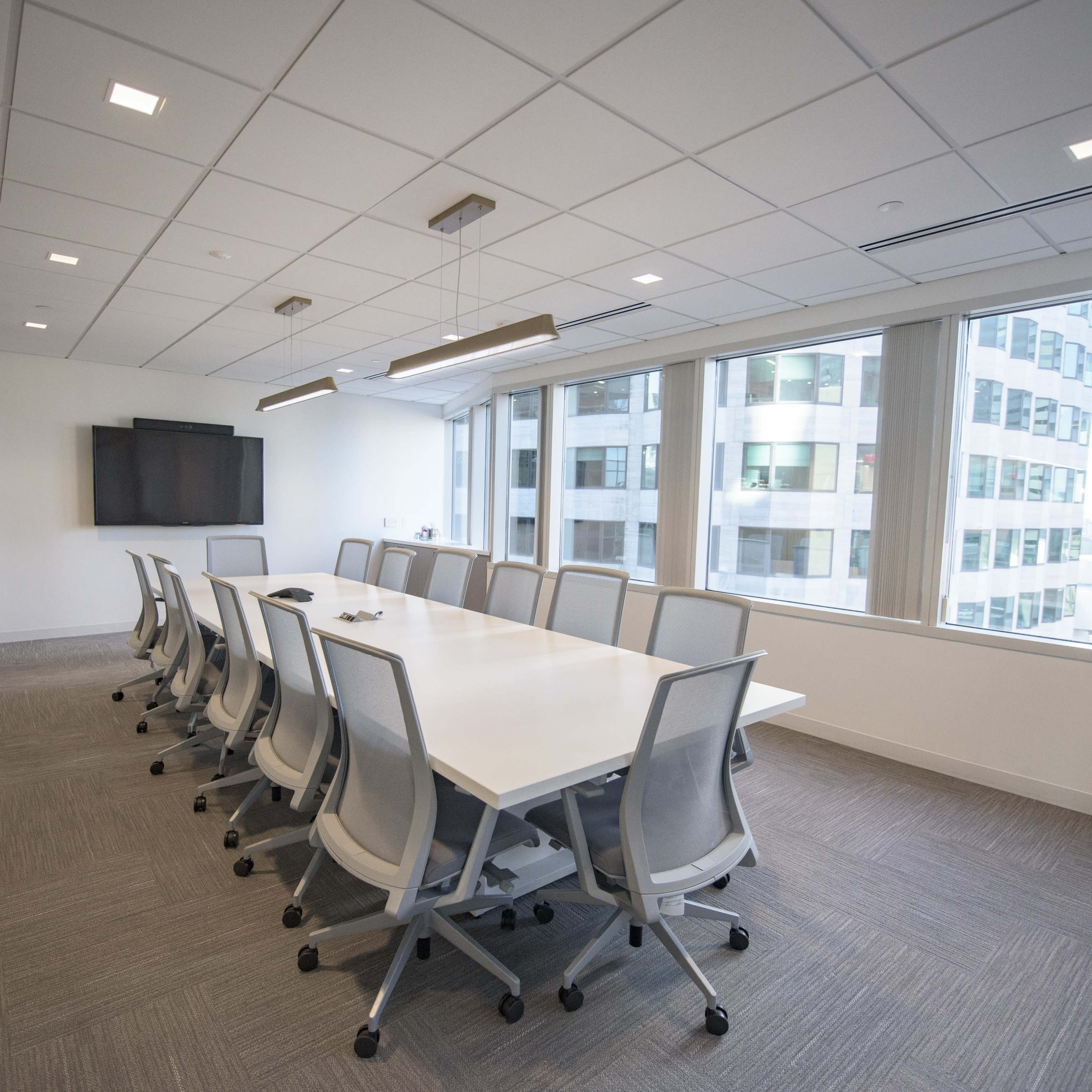 A conference room featuring a long white table, ergonomic chairs, large windows, and a wall-mounted screen.