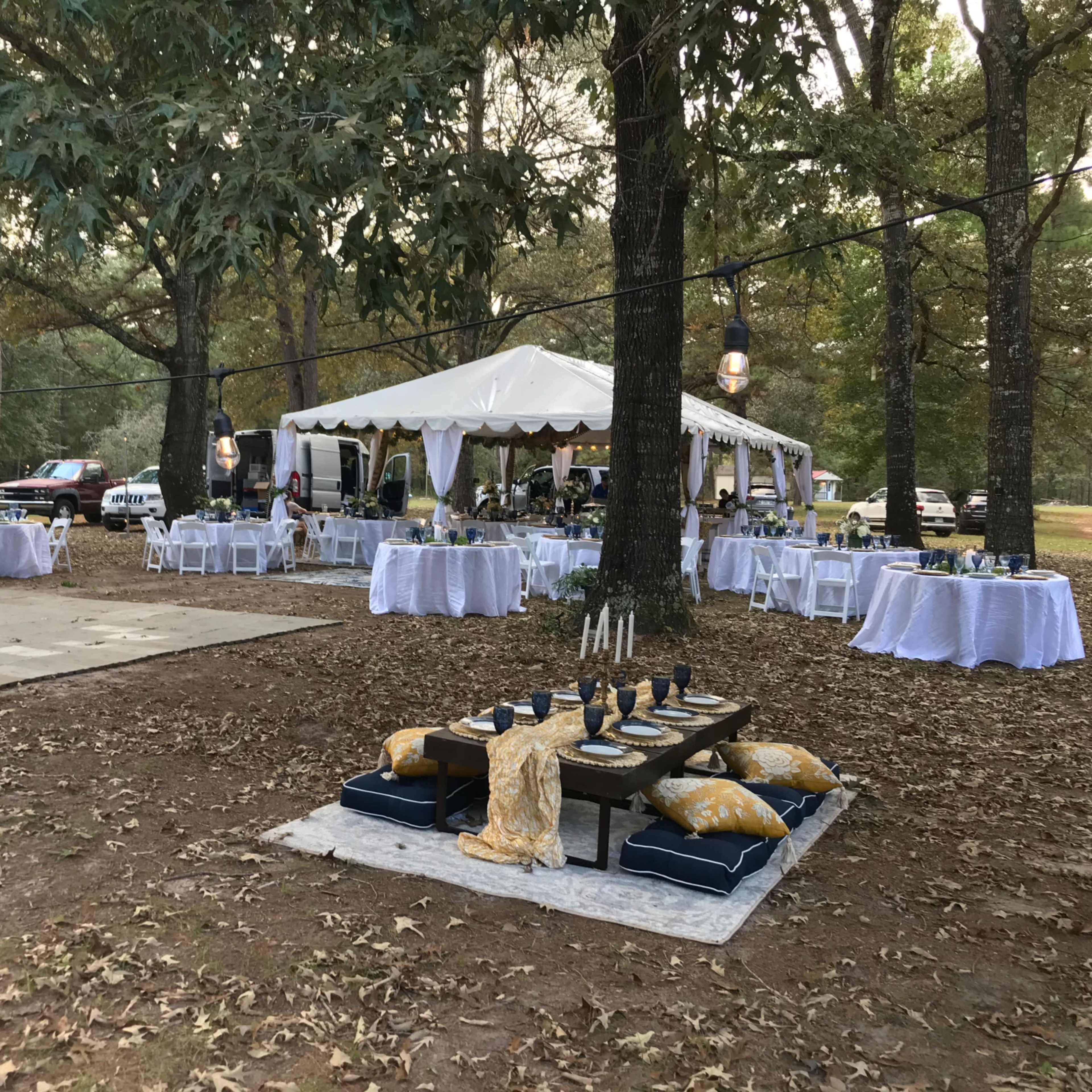 A large outdoor event setup features a tent with tables, chairs, and decorative elements, surrounded by trees and a grassy area.