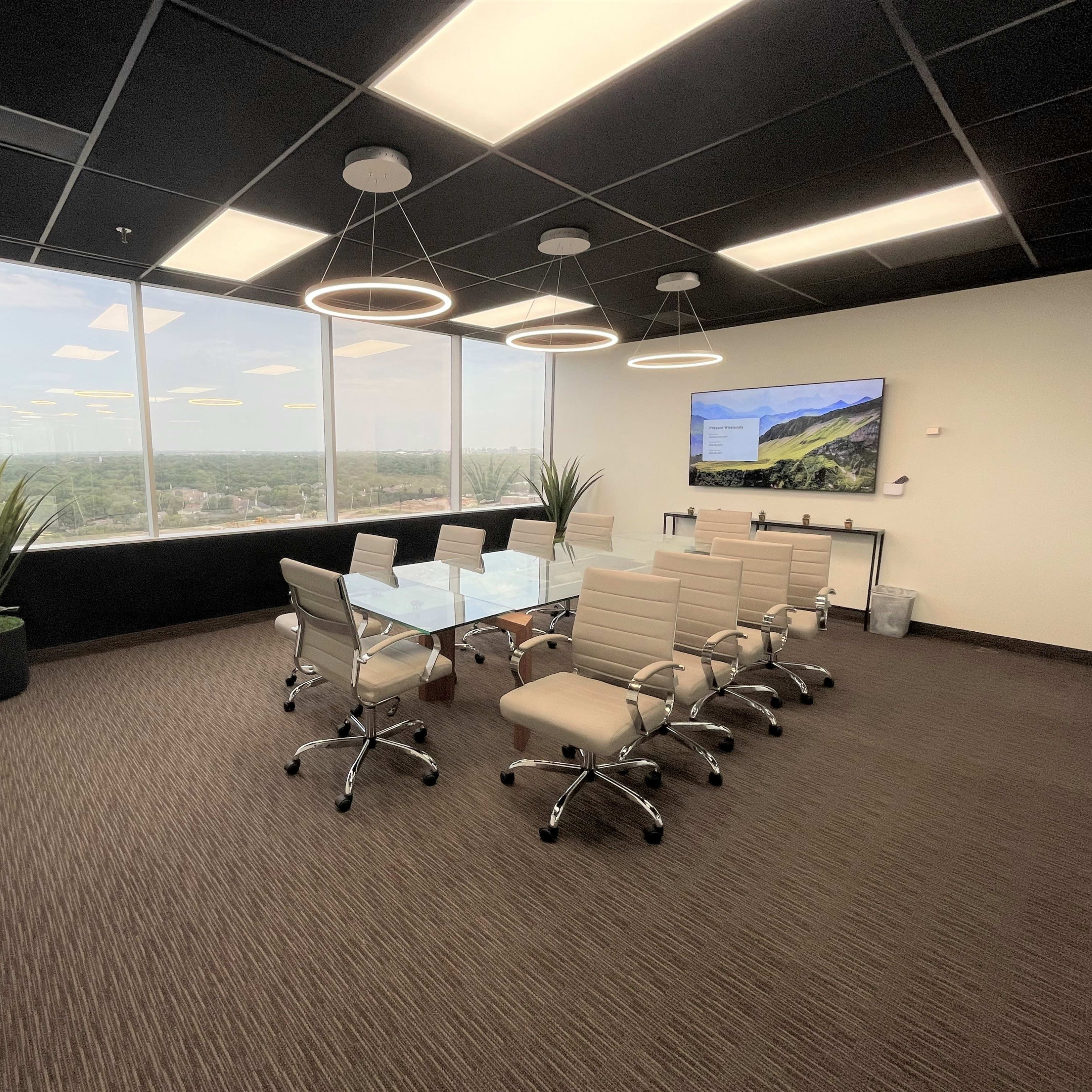 A modern conference room features a glass table surrounded by beige chairs, large windows showcasing a city view, and a screen mounted on the wall.