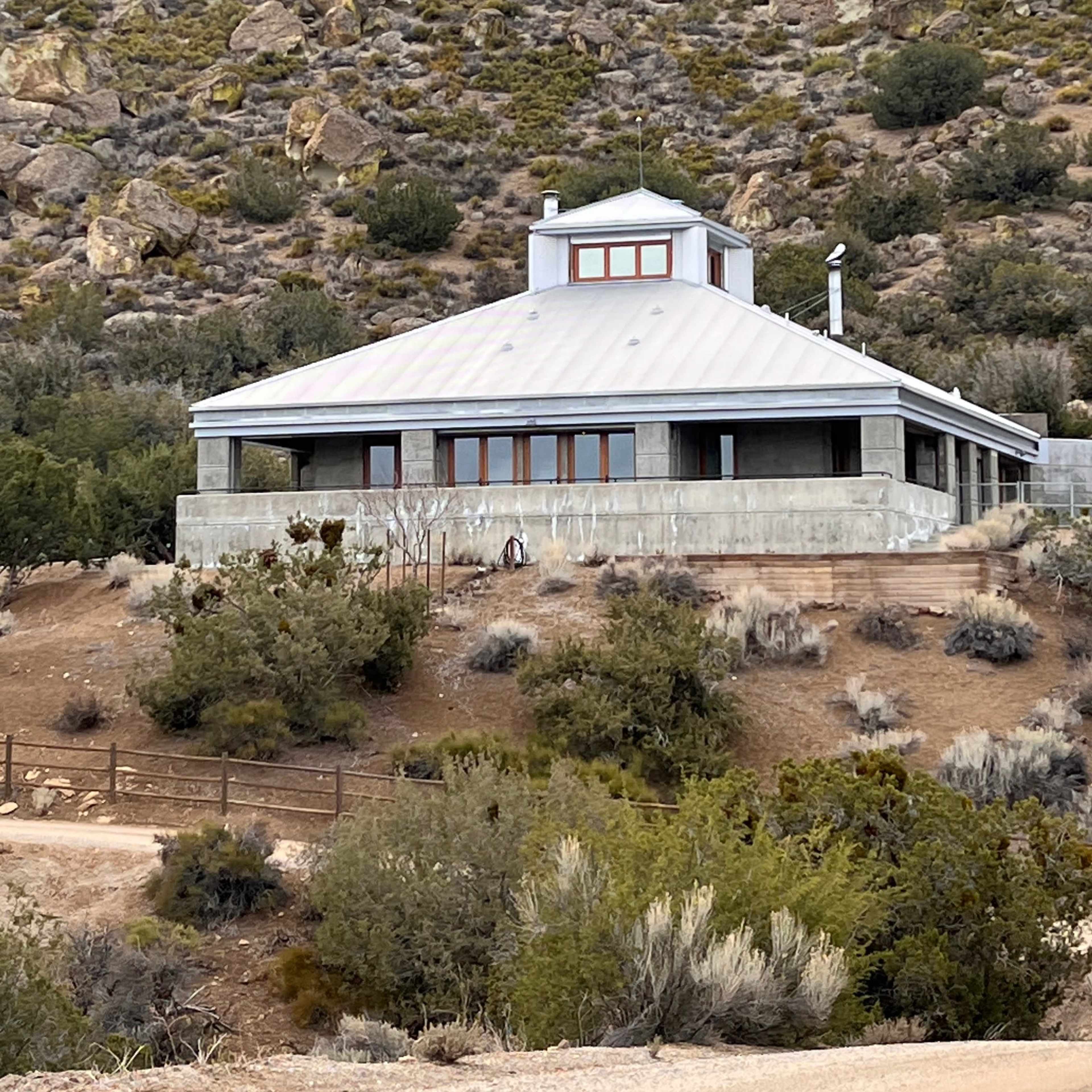 A modern, circular house with large windows is situated on a hillside, surrounded by sparse vegetation and rocky terrain.
