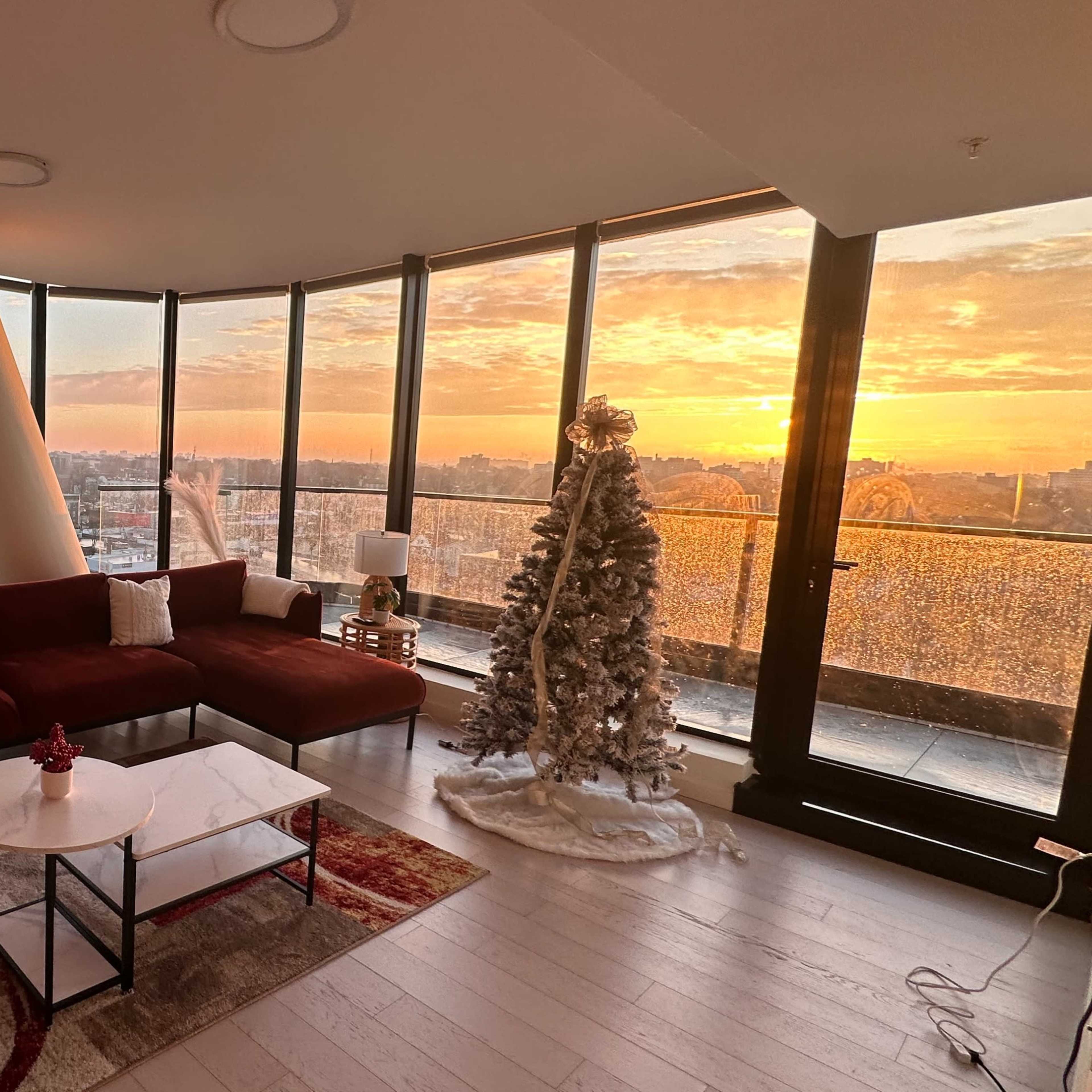 A modern living room with large windows overlooking a sunset, a red sofa, a white coffee table, and a Christmas tree decorated with lights.