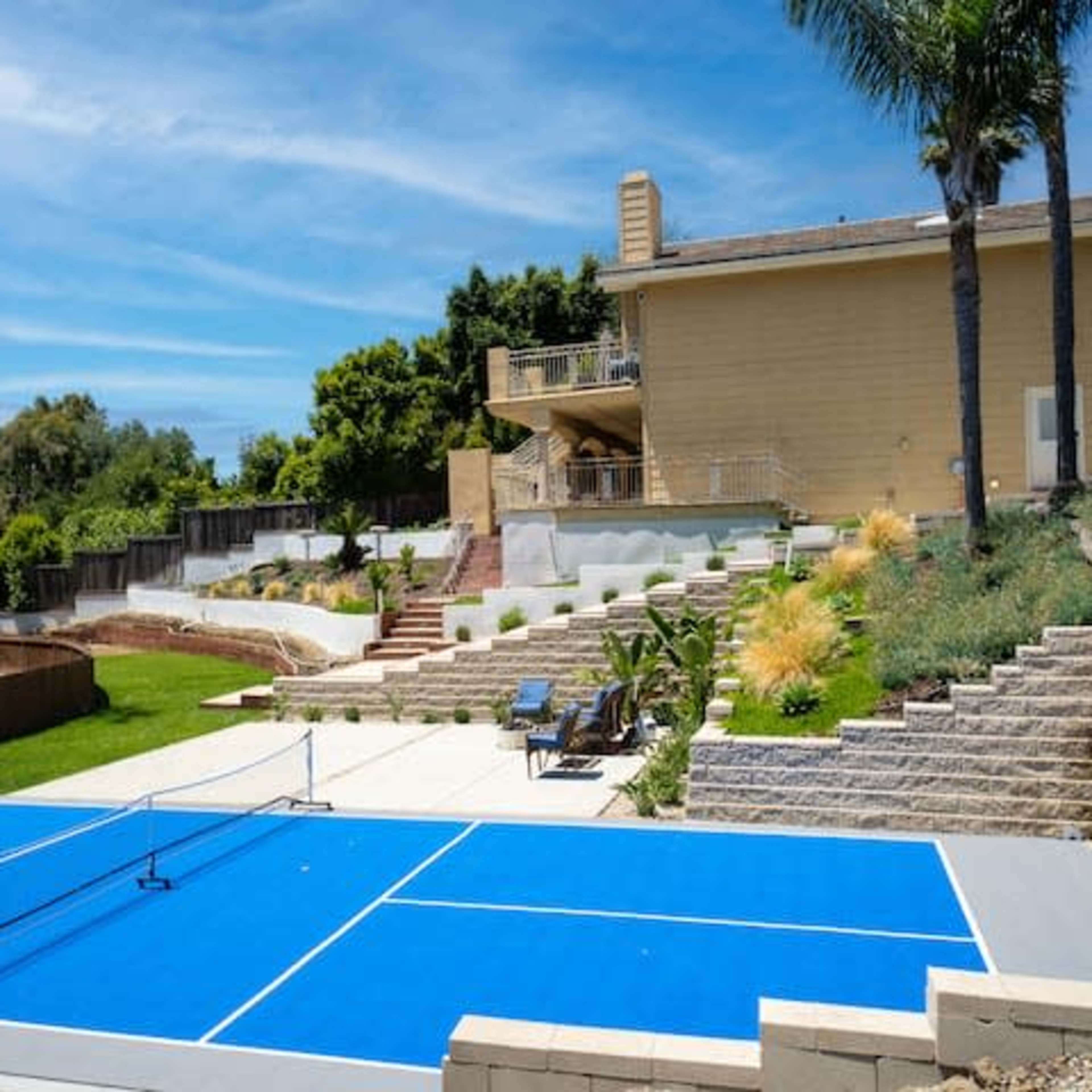 A blue tennis court is situated in a landscaped backyard in front of a two-story house.
