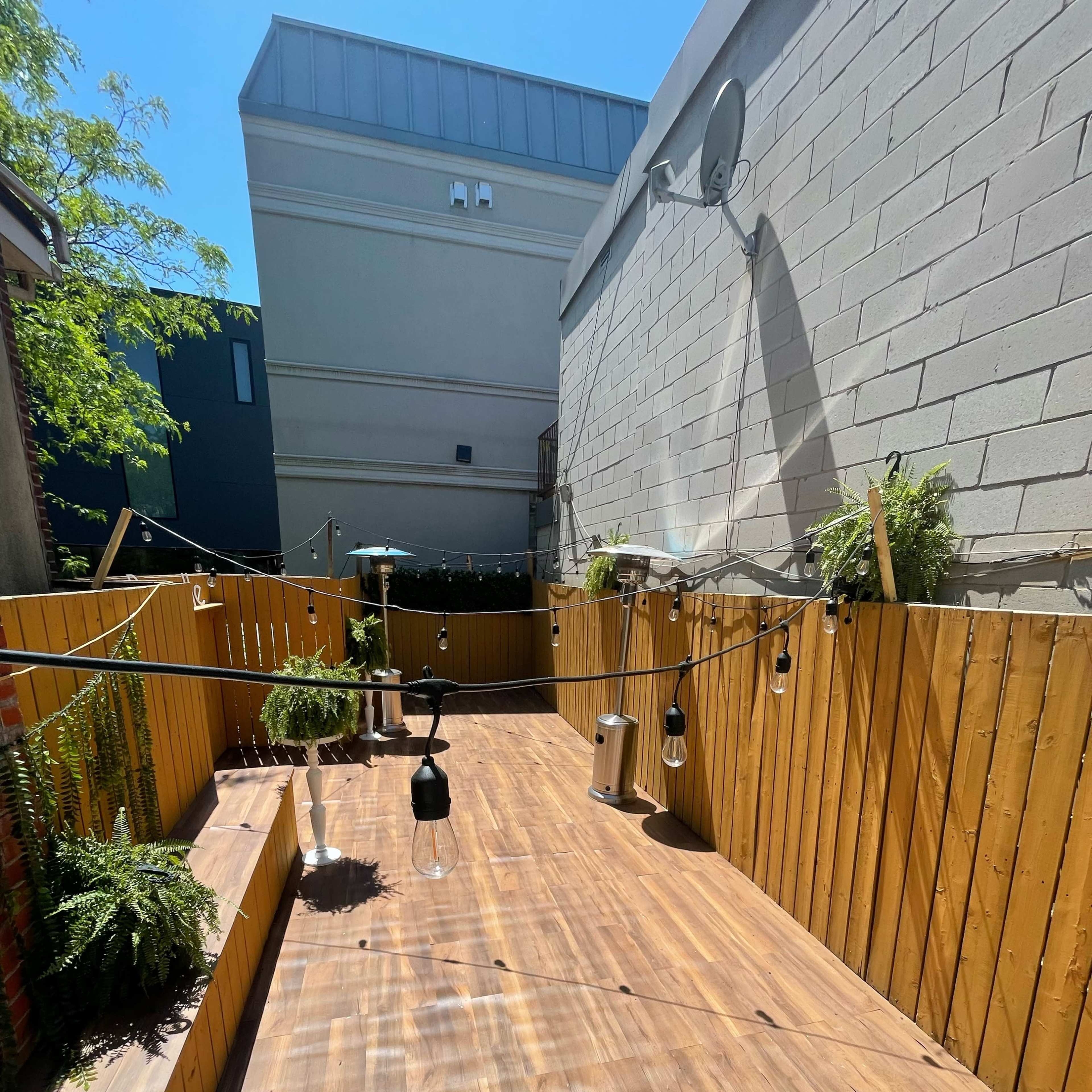 A wooden outdoor space with hanging lights and plants against a backdrop of a brick and gray wall, under a clear blue sky.