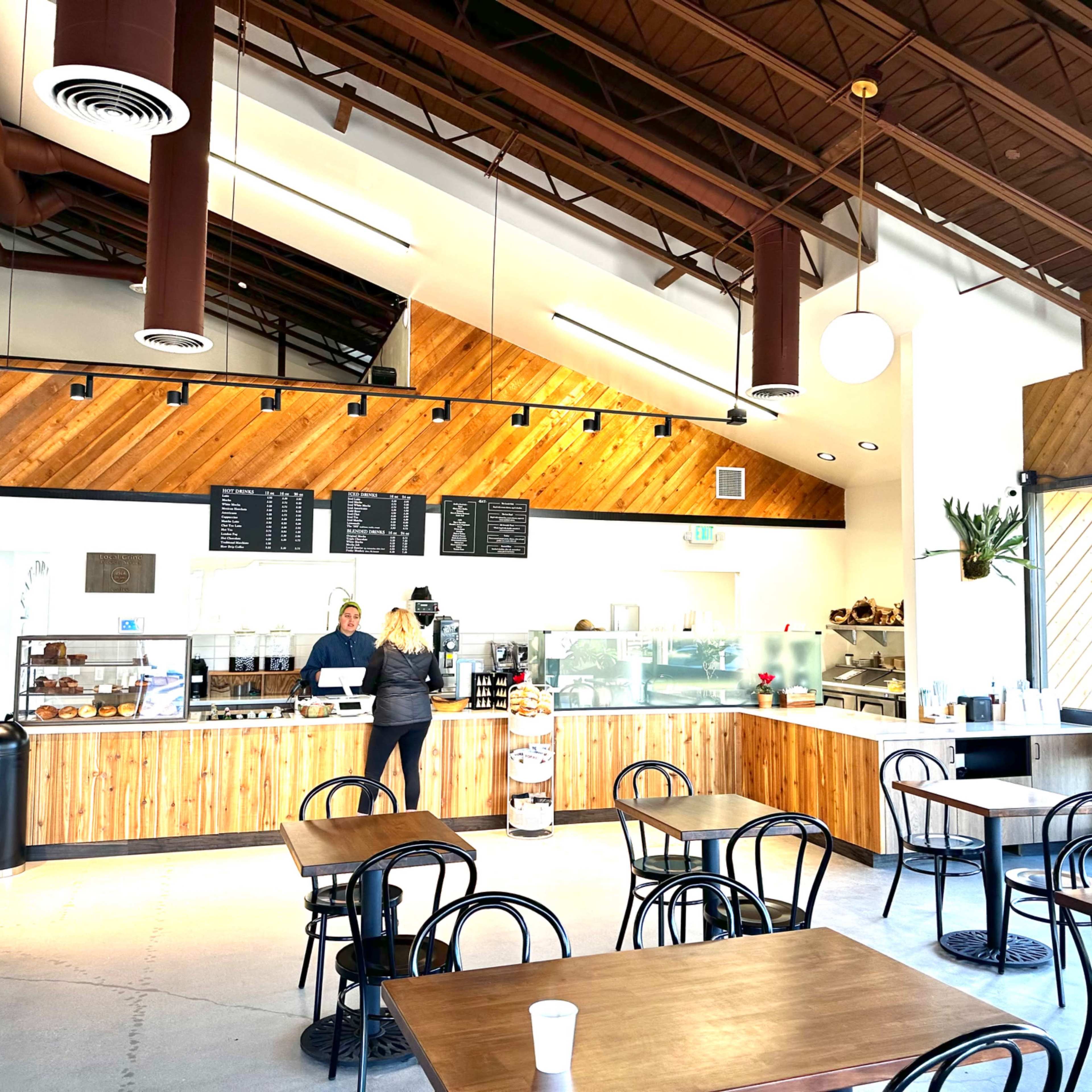A cafe features wooden counter and tables, with a barista attending to customers in a bright, open space with high ceilings and large windows.