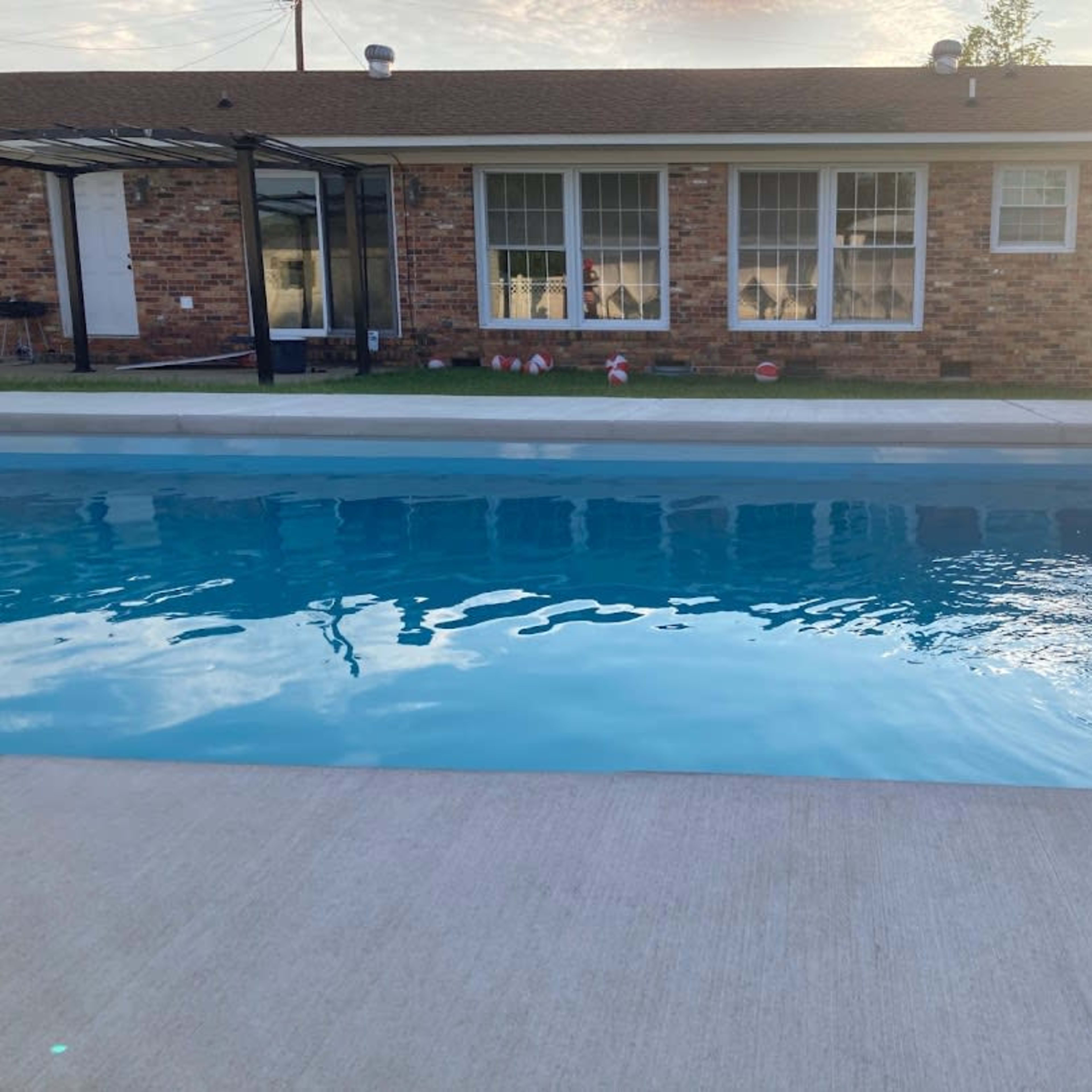 A clear swimming pool with a concrete deck in front of a brick house featuring large windows.