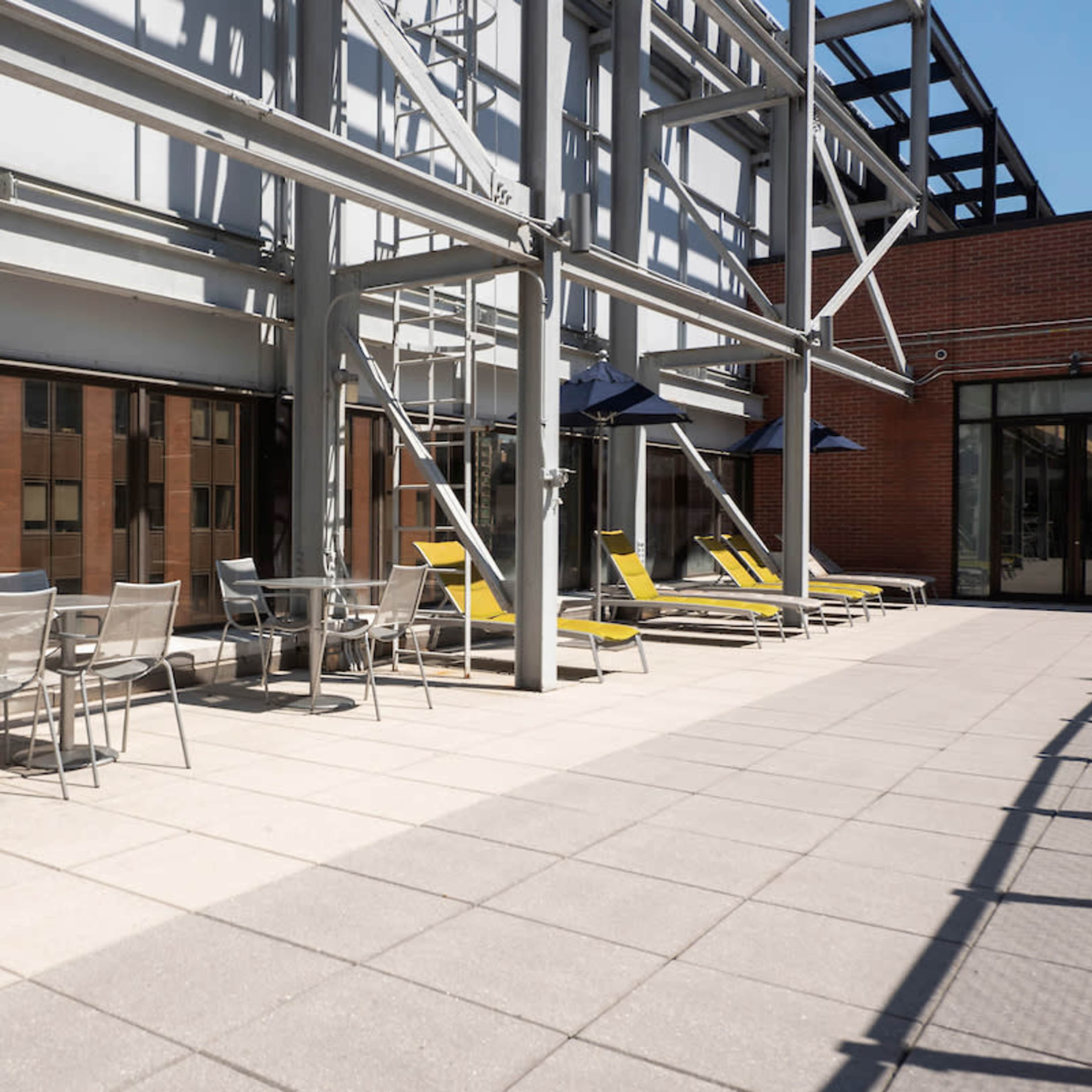 A sunlit rooftop terrace featuring metal furniture, lounge chairs, and umbrellas against a modern building backdrop.