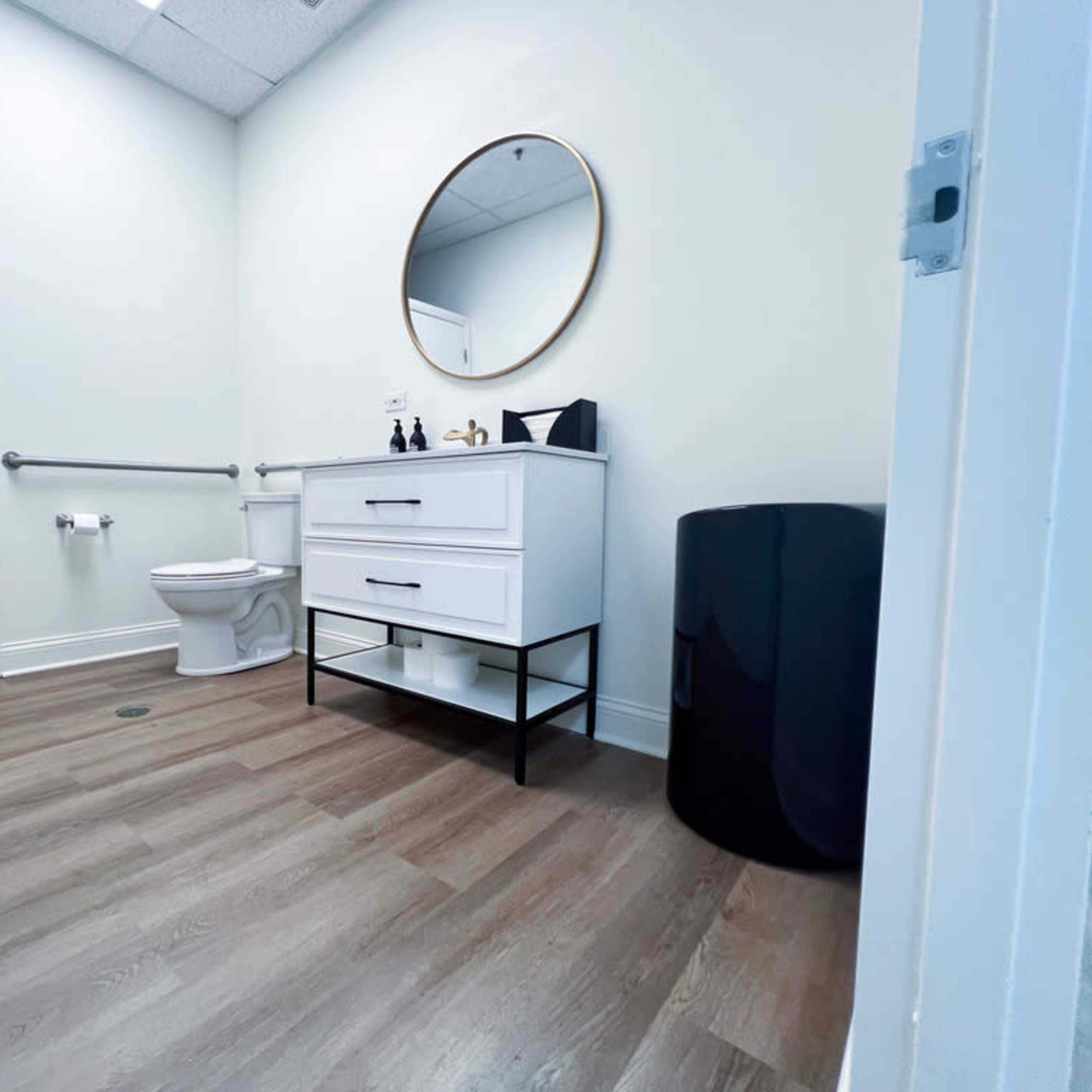 A clean restroom features a white vanity with a round mirror, a toilet, and a black wastebasket against light-colored walls and wooden flooring.