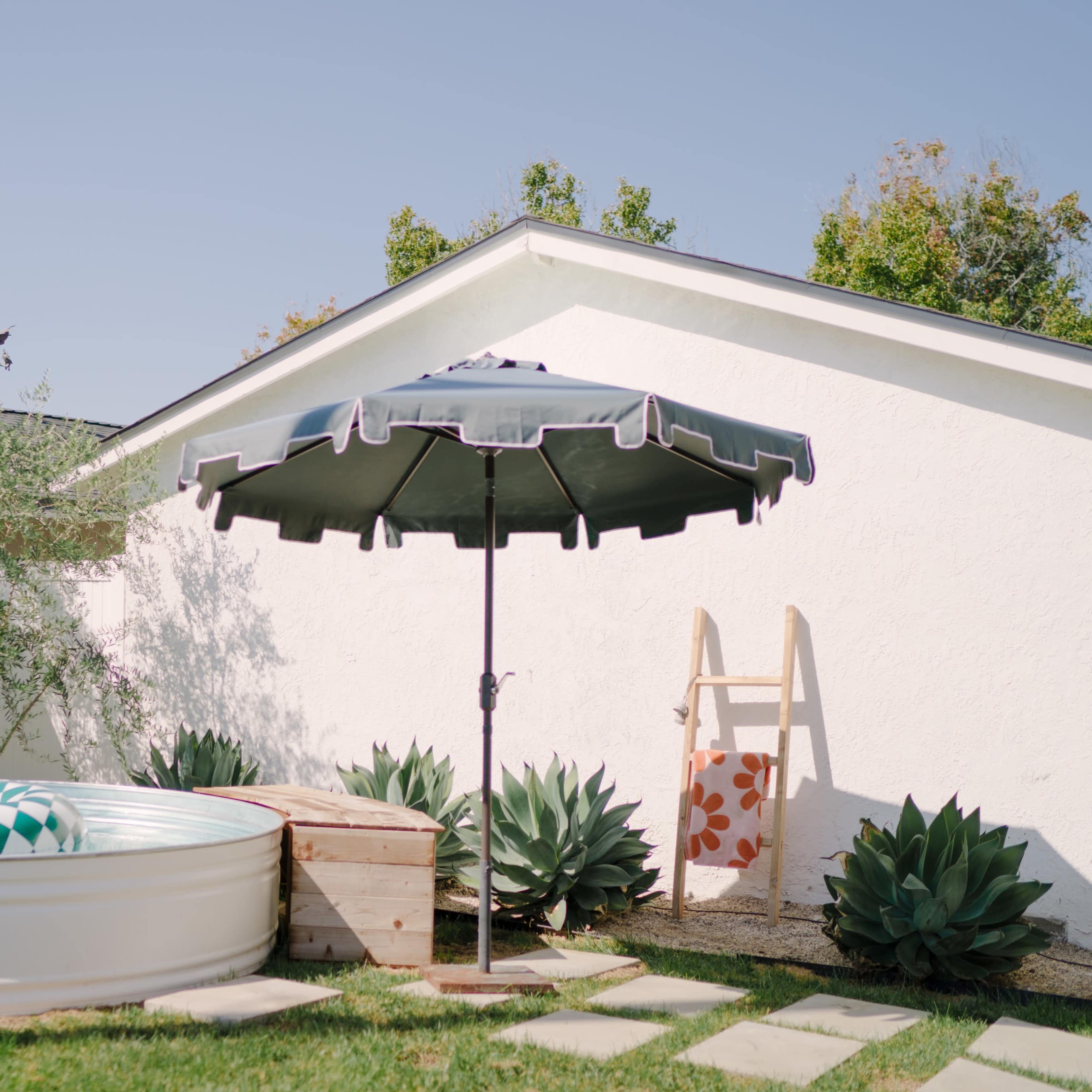 A small above-ground pool is situated in a yard with an umbrella and a ladder, surrounded by greenery and decorative plants.