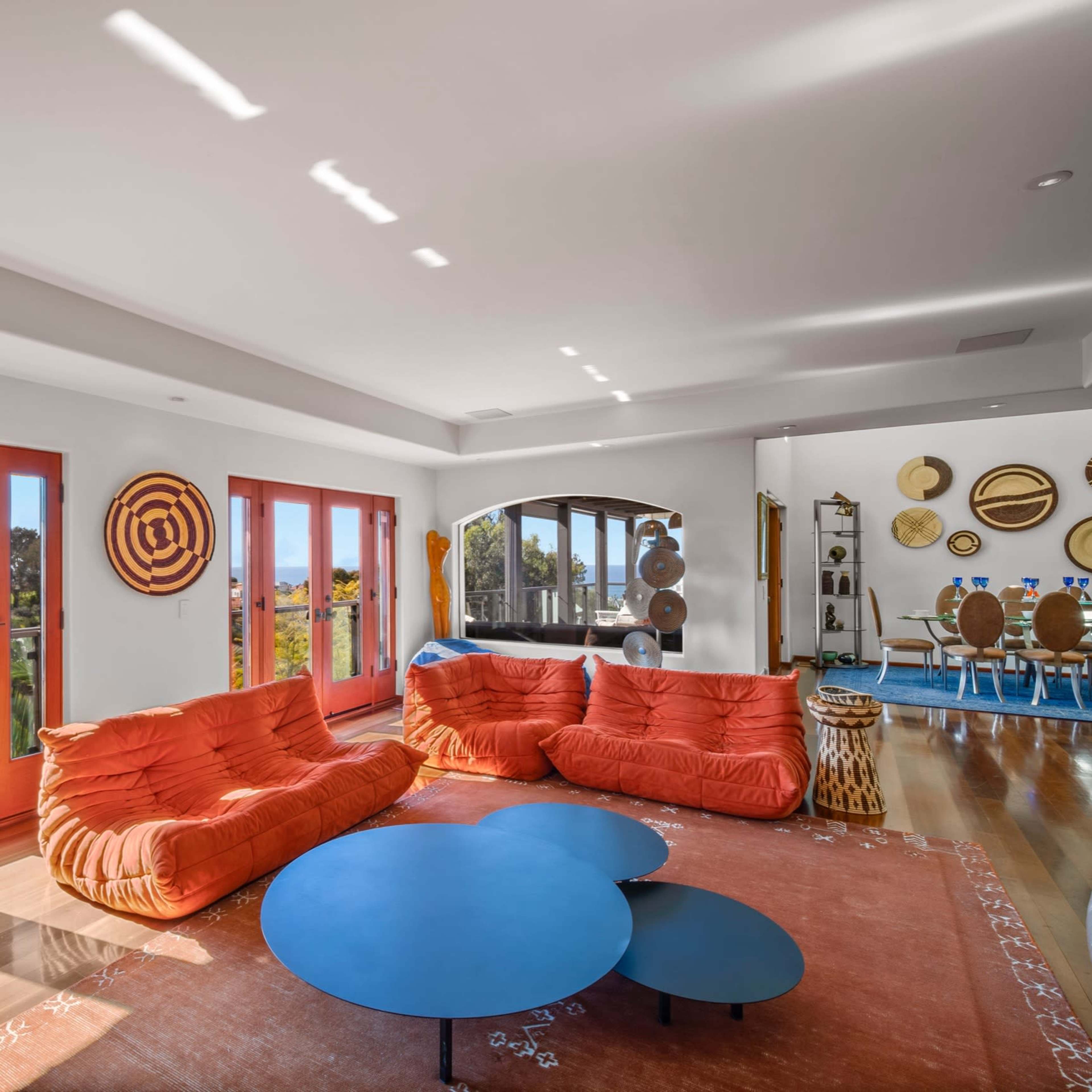 A bright living room features orange sofas, a round blue coffee table, and large windows opening to a scenic view.