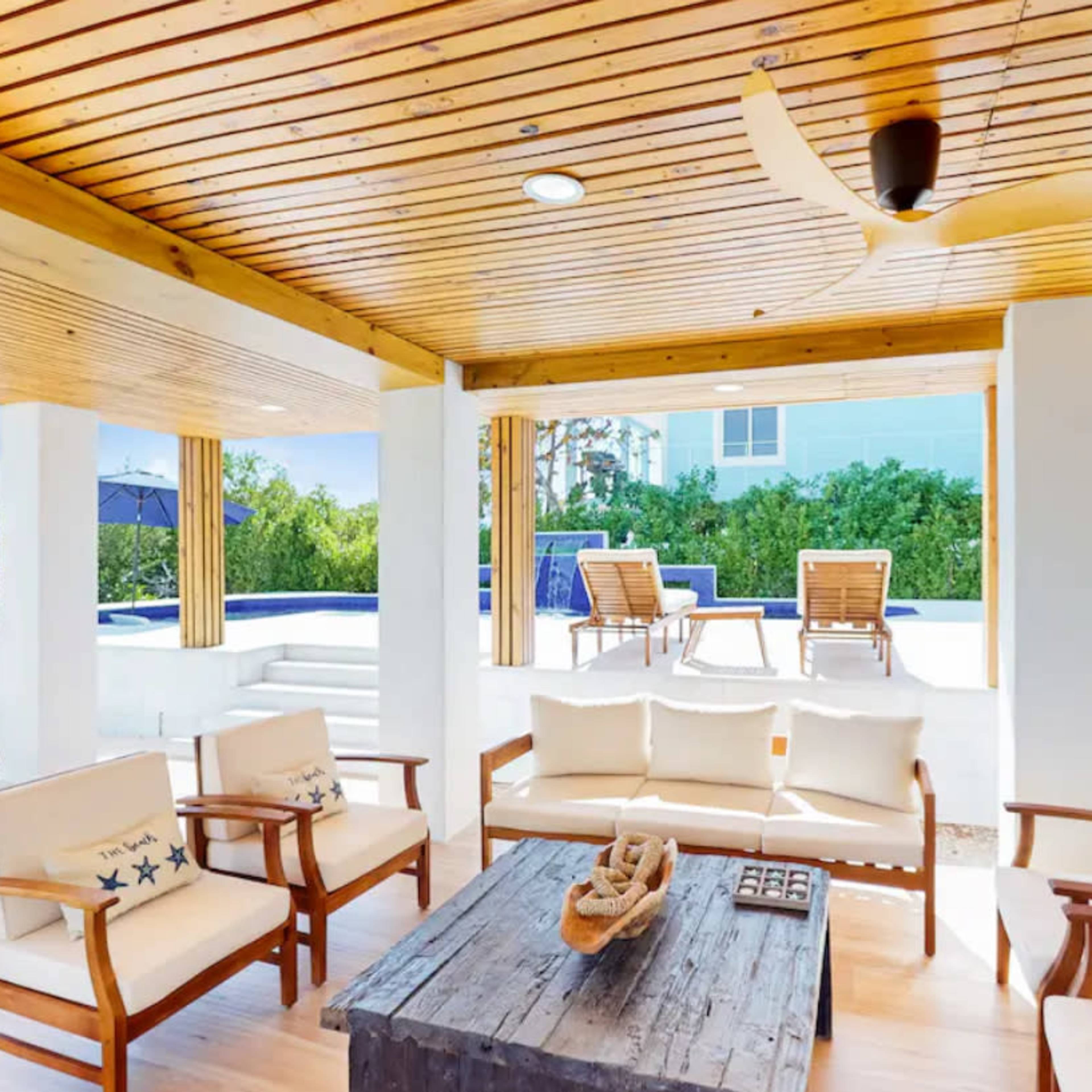 A covered outdoor seating area features wooden furniture and a coffee table, with a view of a pool and lounge chairs in the background.