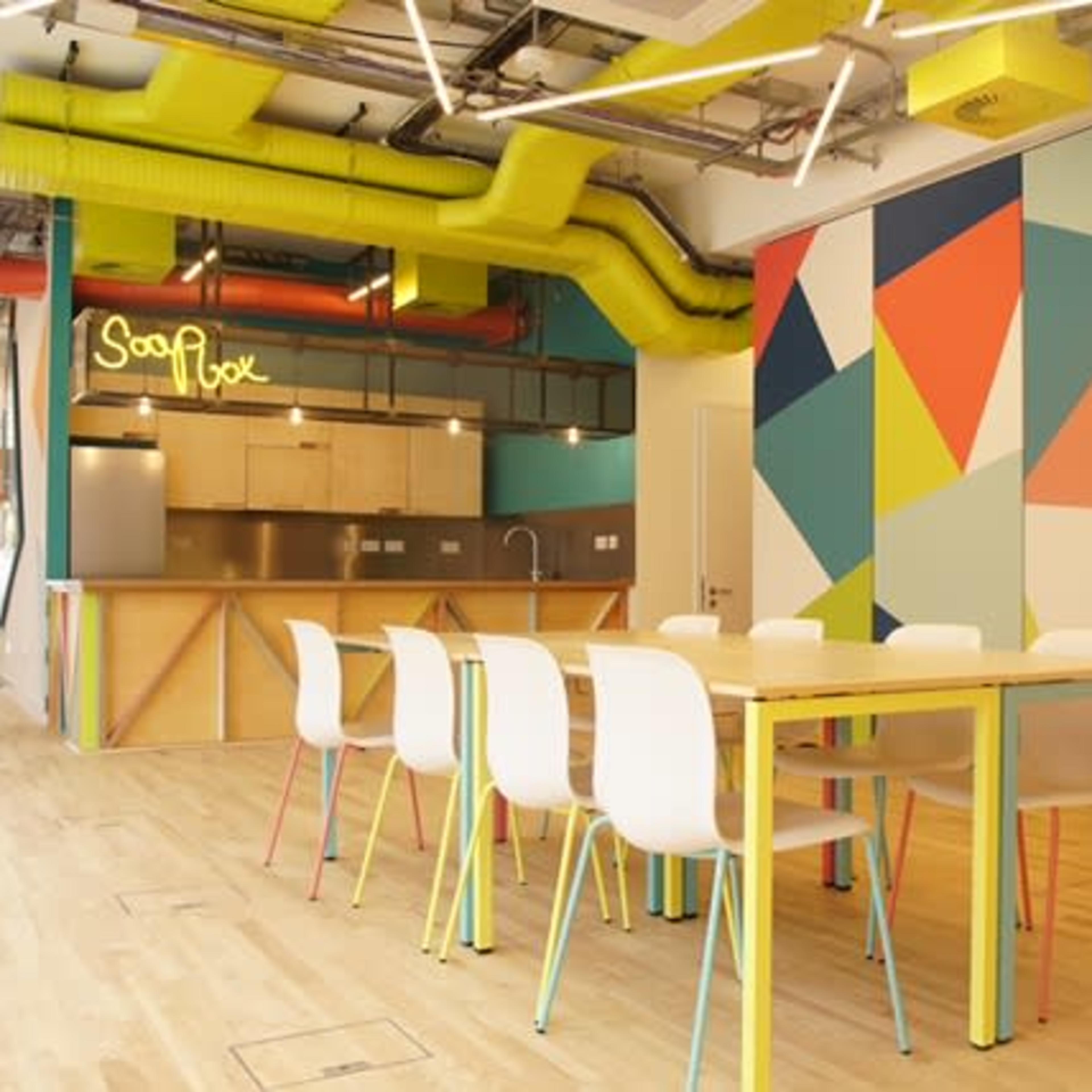 A modern kitchen and dining area featuring a long table with white chairs, colorful geometric wall designs, and a bright green ventilation system.