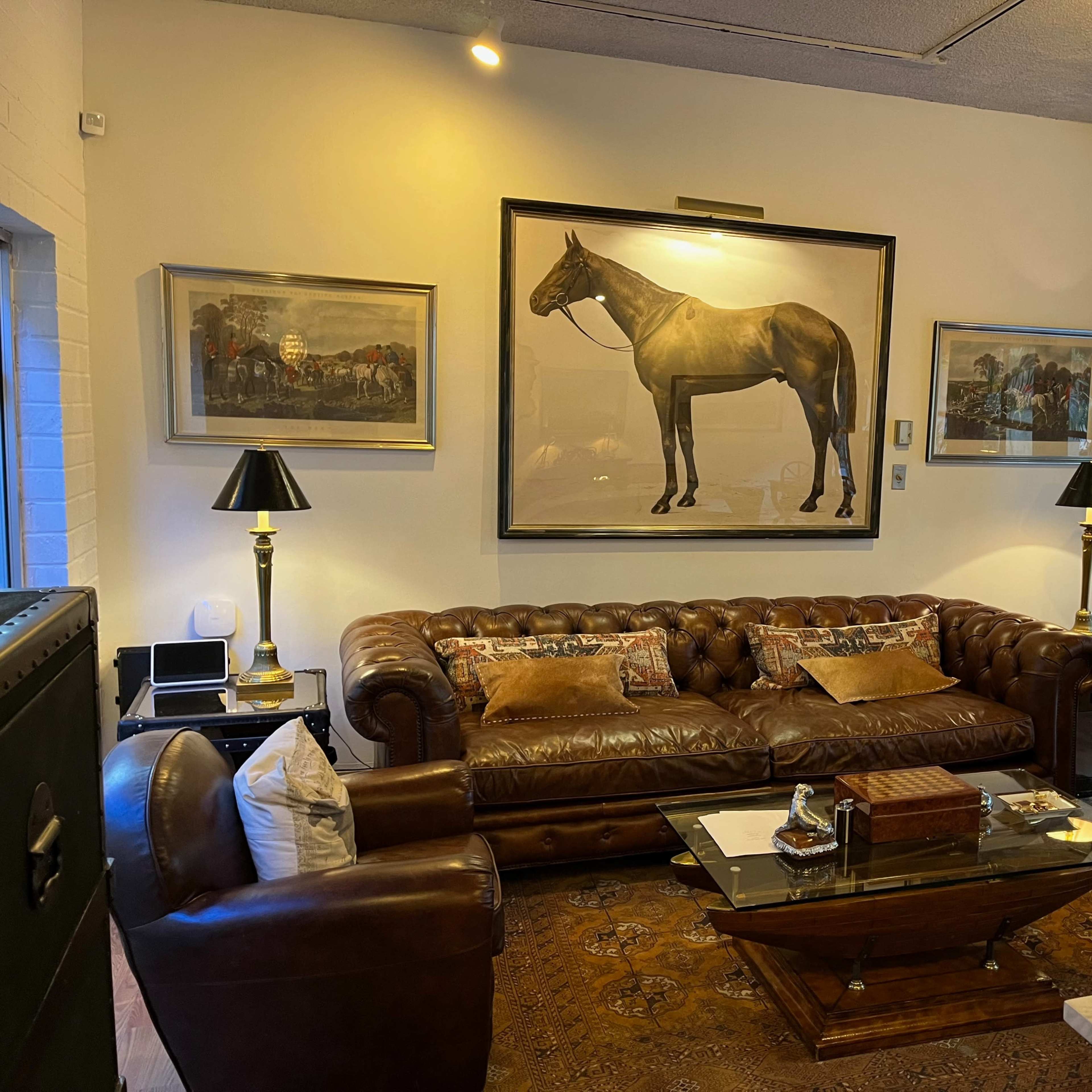 A living room featuring a brown leather sofa, a glass coffee table, and a large framed painting of a horse on the wall.