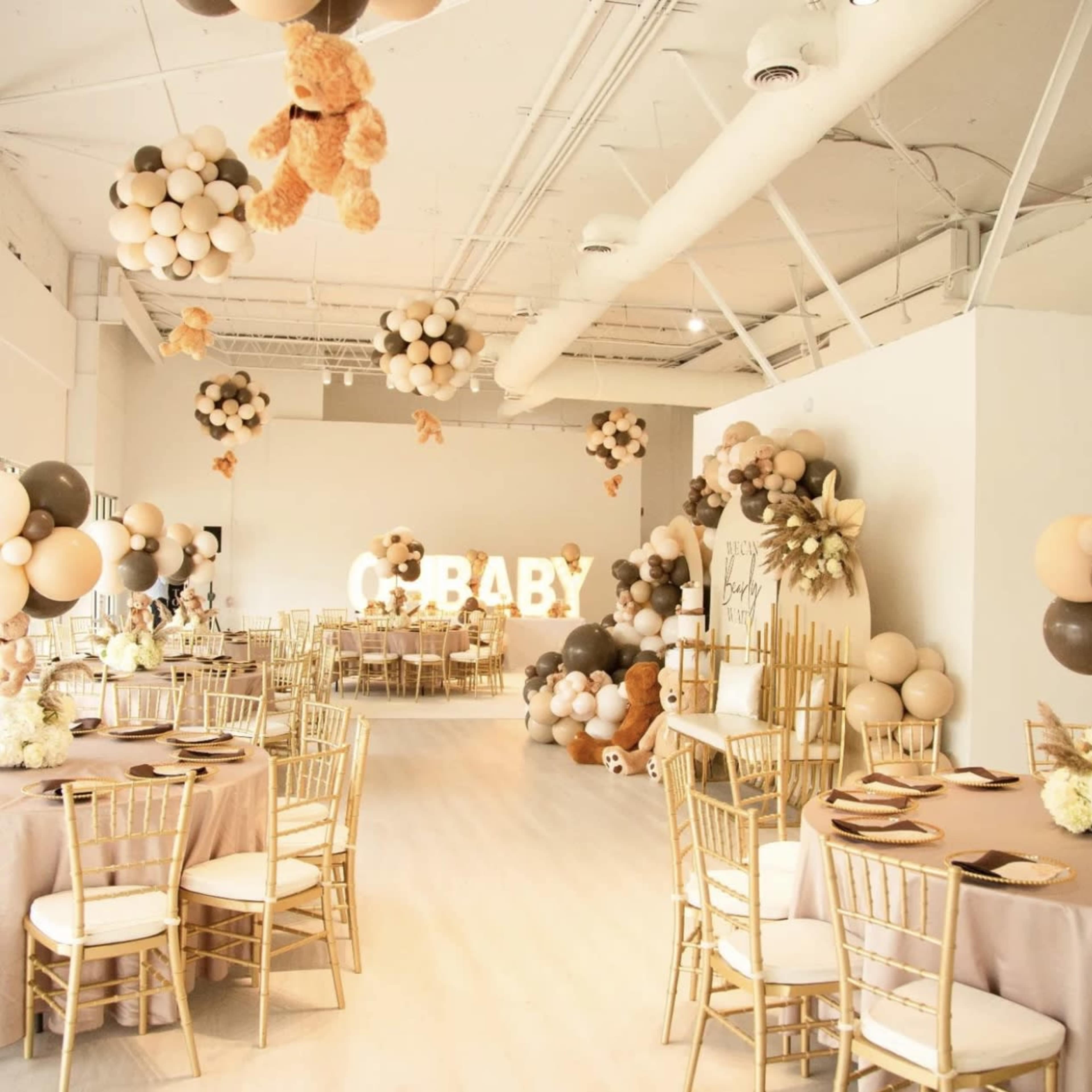 A celebratory room decorated for a baby shower, featuring tables with gold chairs, balloons in various colors, and a "BABY" light display.