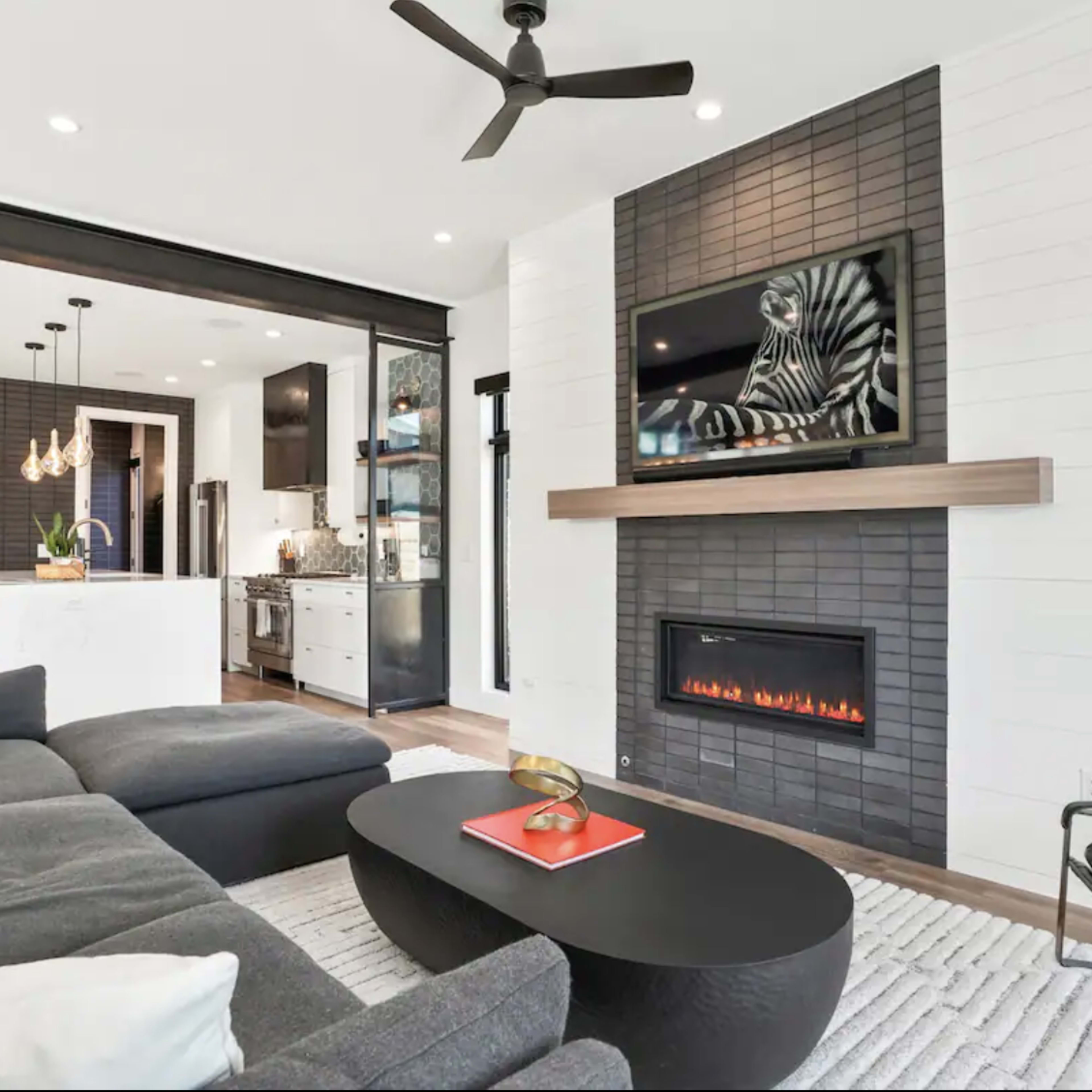 A modern living room with a sectional sofa, an elegant coffee table, a wall-mounted television above a sleek fireplace, and an open view of an adjacent kitchen.