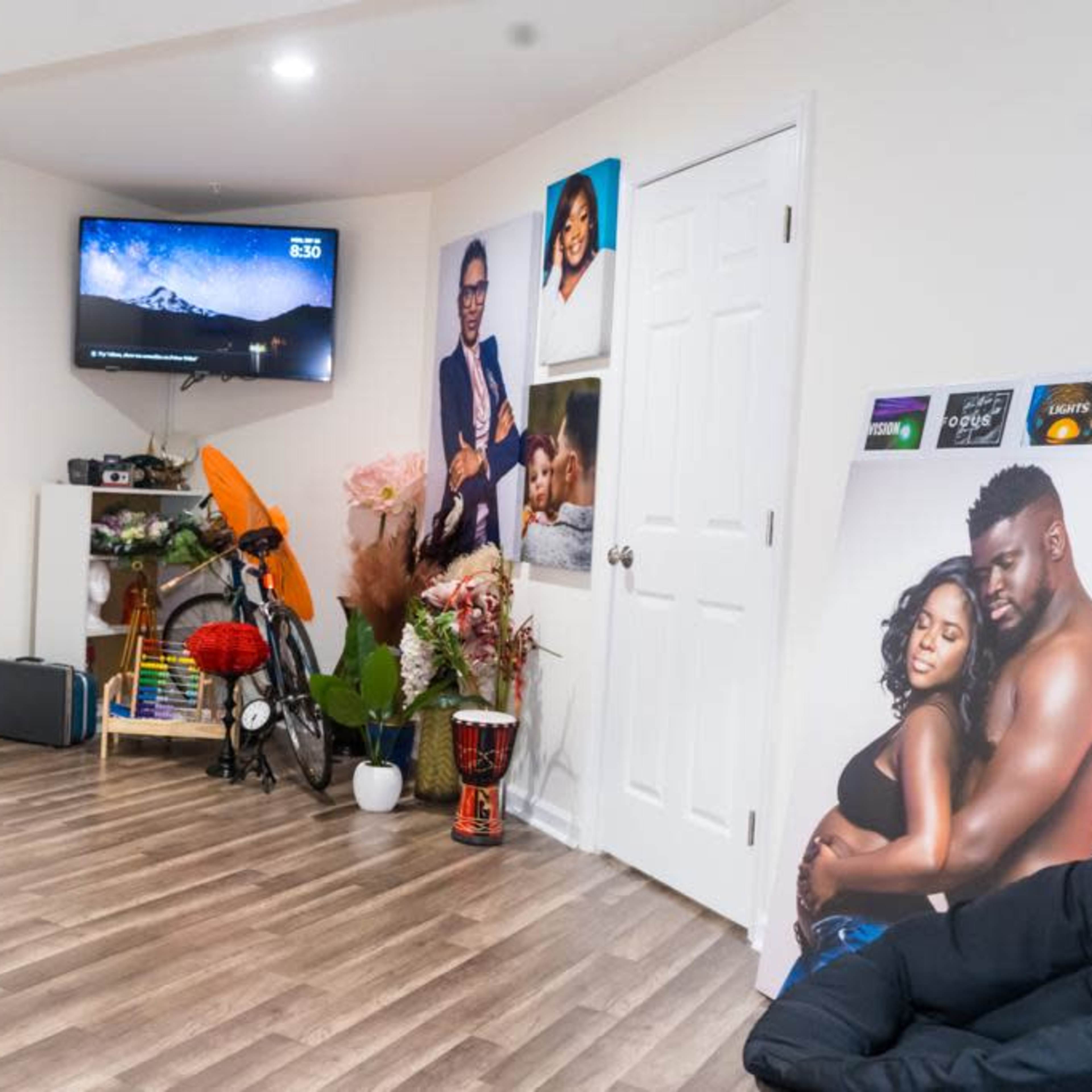 A room with hardwood flooring, featuring a blue bicycle, a television mounted on the wall, a variety of plants, and several wall decorations, including posters and shelves.