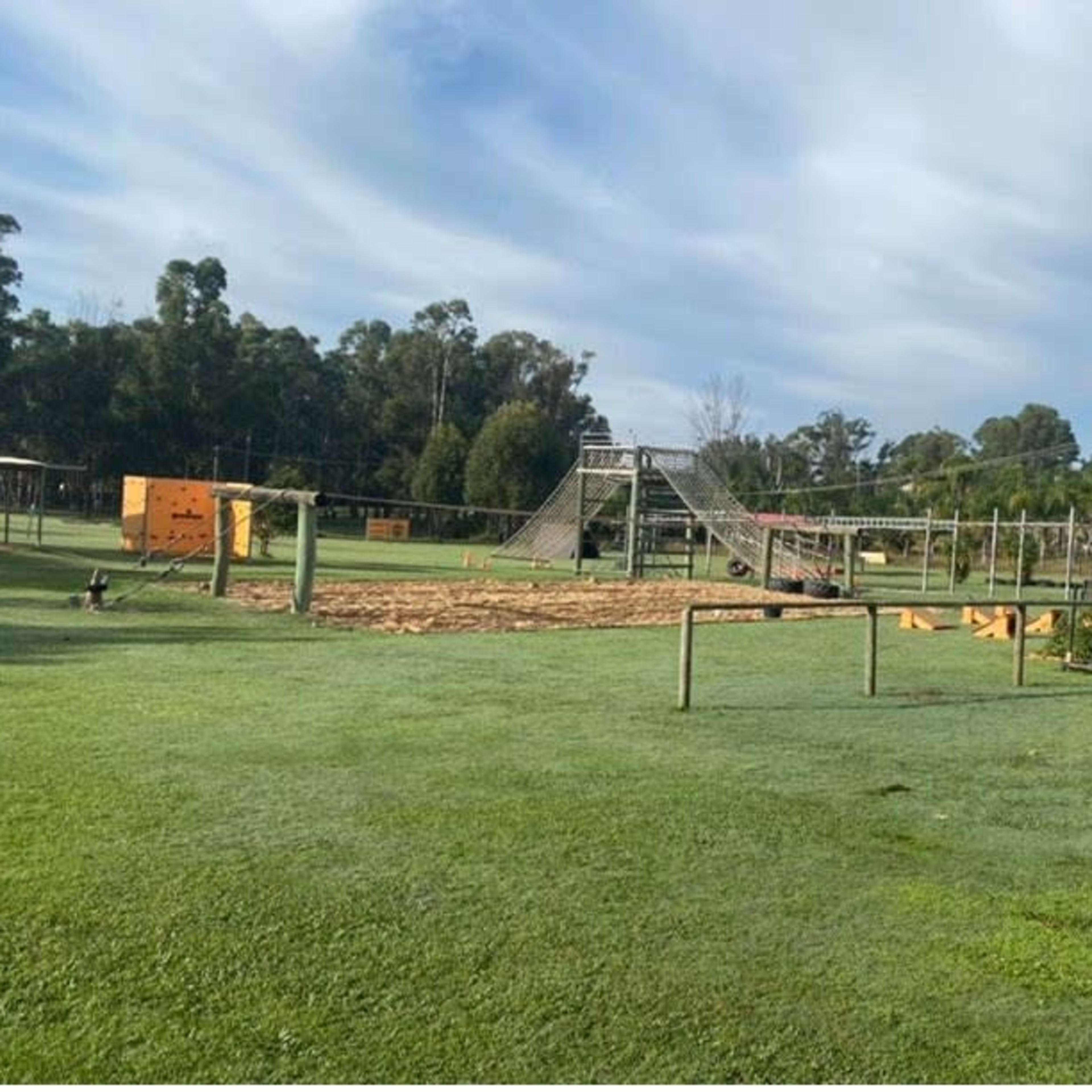 A playground with various climbing structures and equipment set on a grassy field, surrounded by trees.