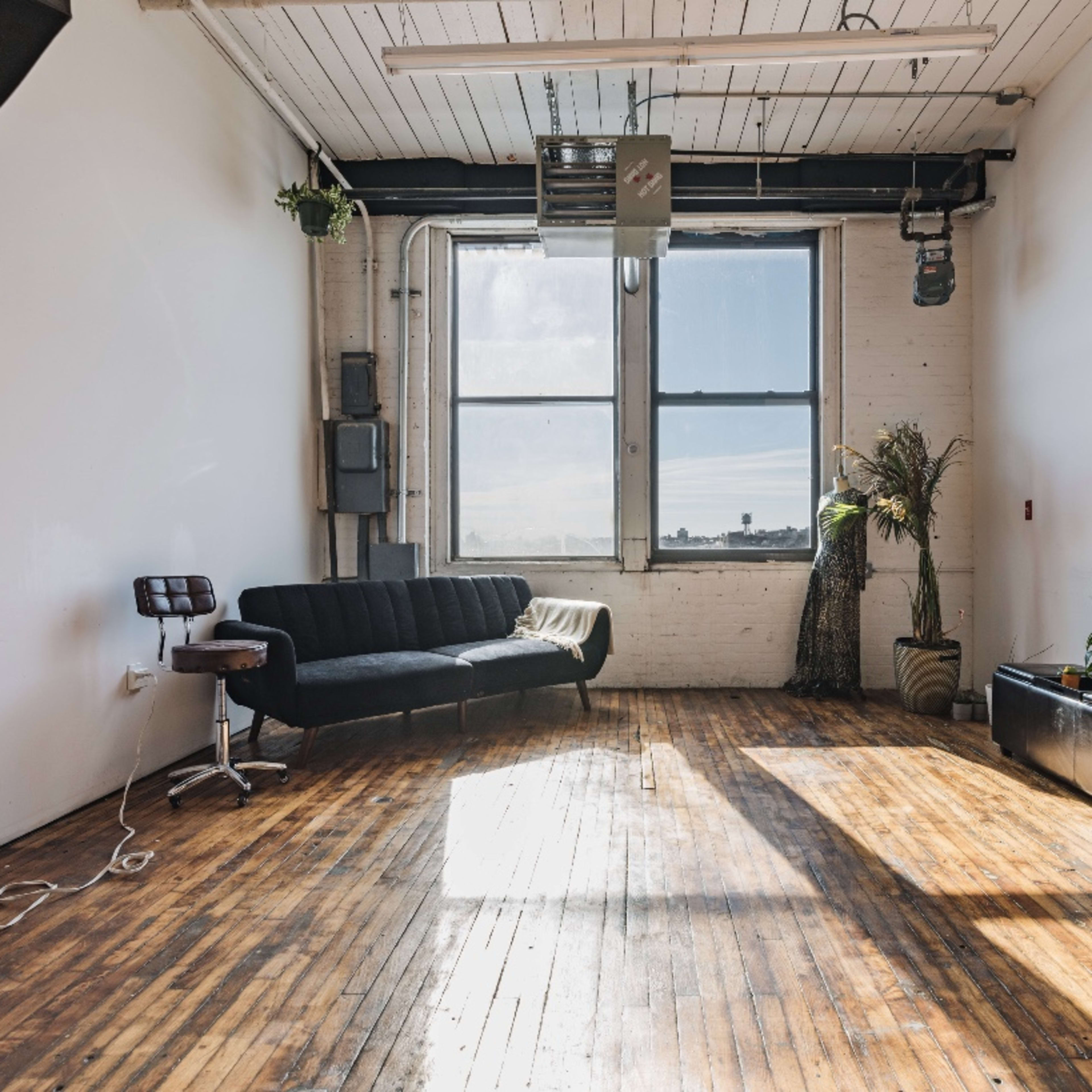 A spacious, empty room with wooden floors, a large window allowing natural light, a black sofa, and a couple of potted plants.